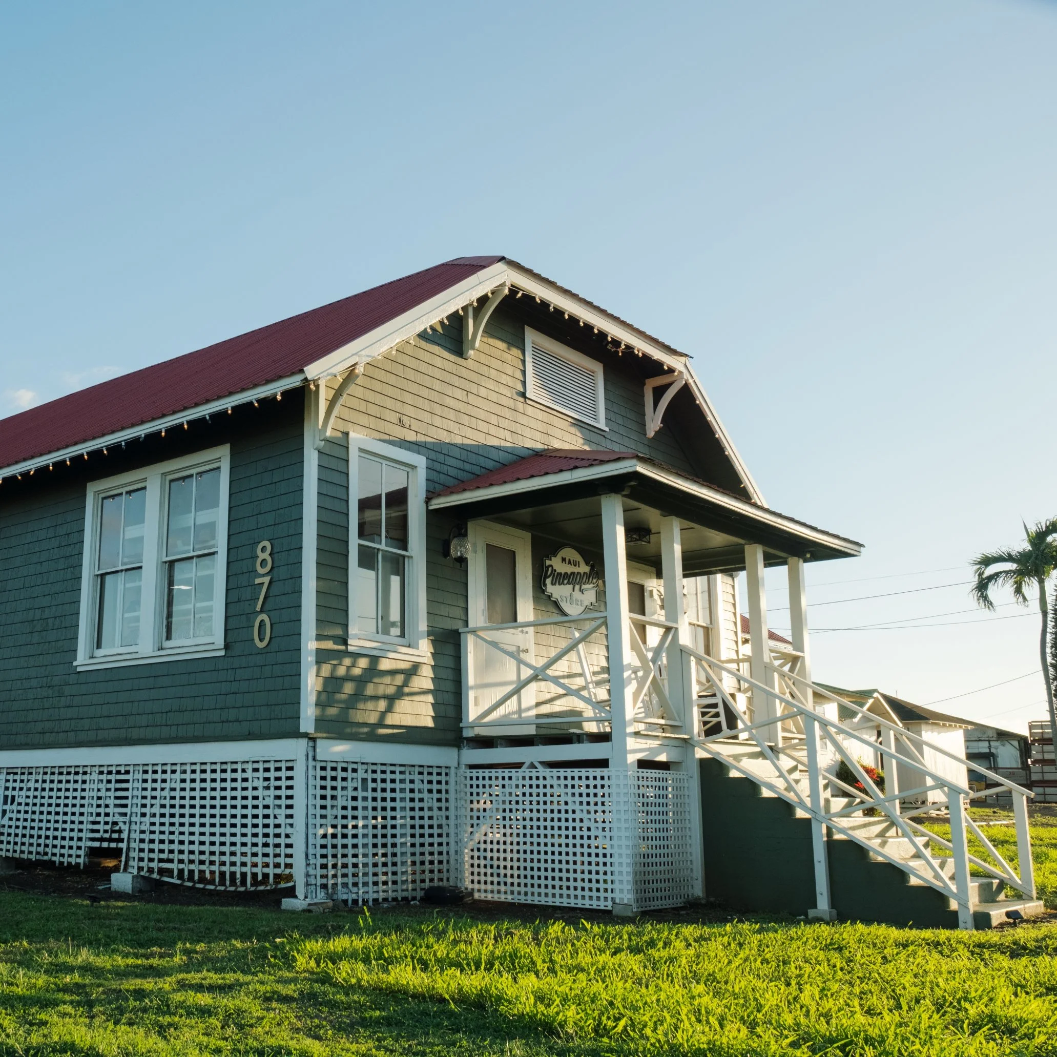 A house with light green siding, a red roof, white trim, and a front porch decorated with string lights. The house has white-framed windows and a sign reading 'Maui Pineapple Store' near the porch. There is a grassy yard in front and a palm tree to the right, with a clear blue sky overhead.