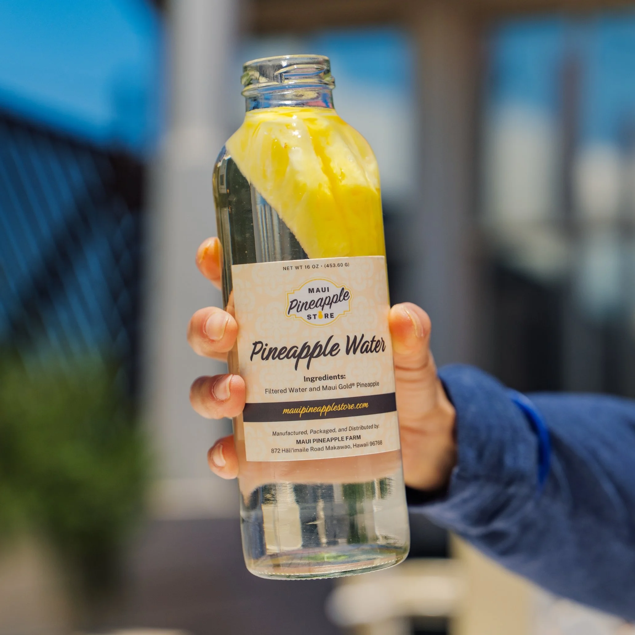 A small glass bottle of Maui Pineapple Water with a label showing ingredients and branding. The bottle is held by a person's hand with a blurred background.