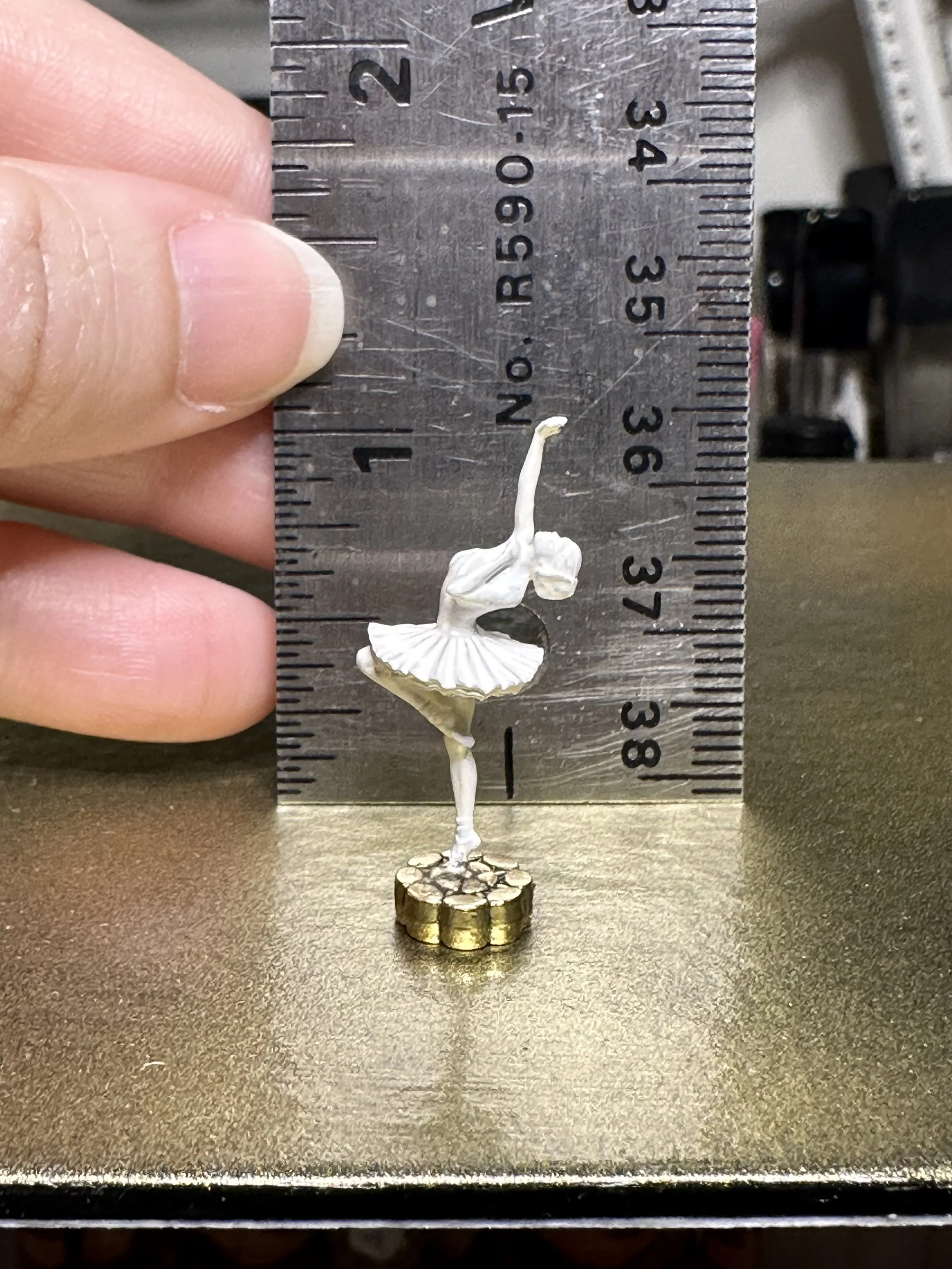 Miniature ballet dancer figurine, approximately 3 inches tall, with a gold-colored base, positioned against a ruler showing height, held near a person's thumb and index finger.