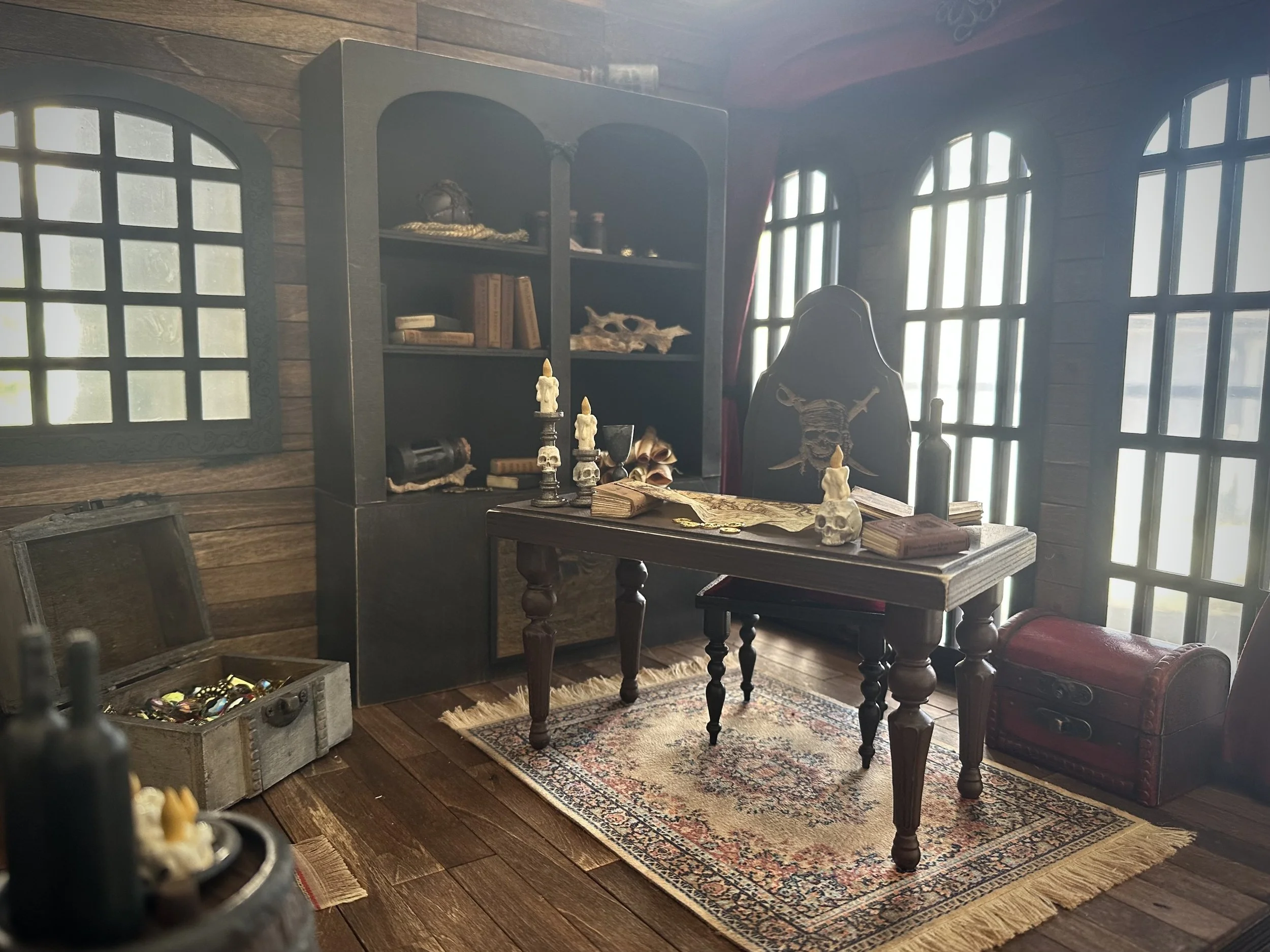 A pirate's captain's quarters with wooden walls and floor, featuring a wooden desk with books and skulls, a bookshelf with artifacts, a red chest, and a writing chair with a skull pillow. Large arched windows let in natural light.