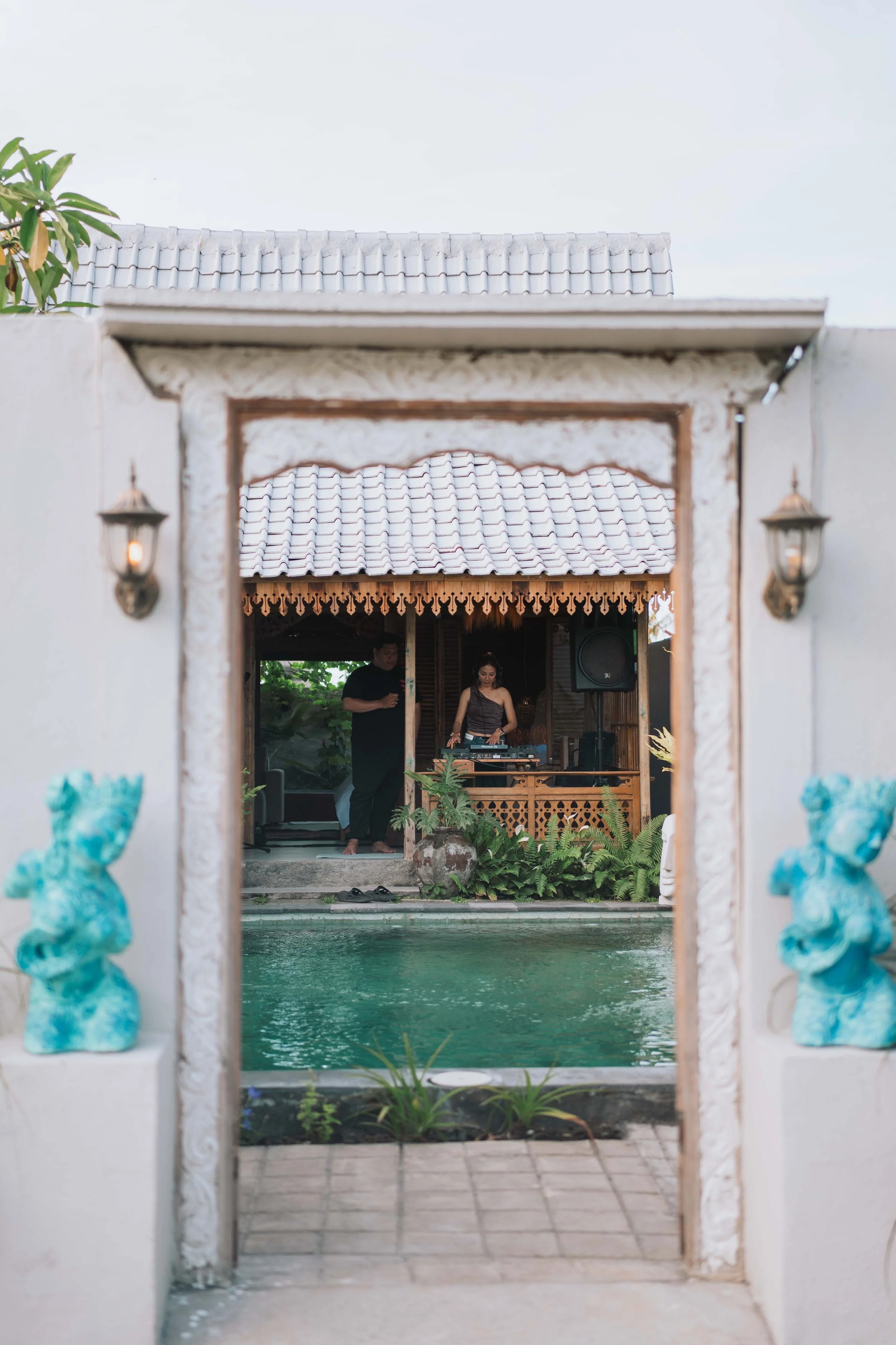View through a decorative gate of a backyard with a swimming pool, tropical plants, and a DJ playing music with speakers in a wooden pavilion, two people, one holding a drink, in the background.