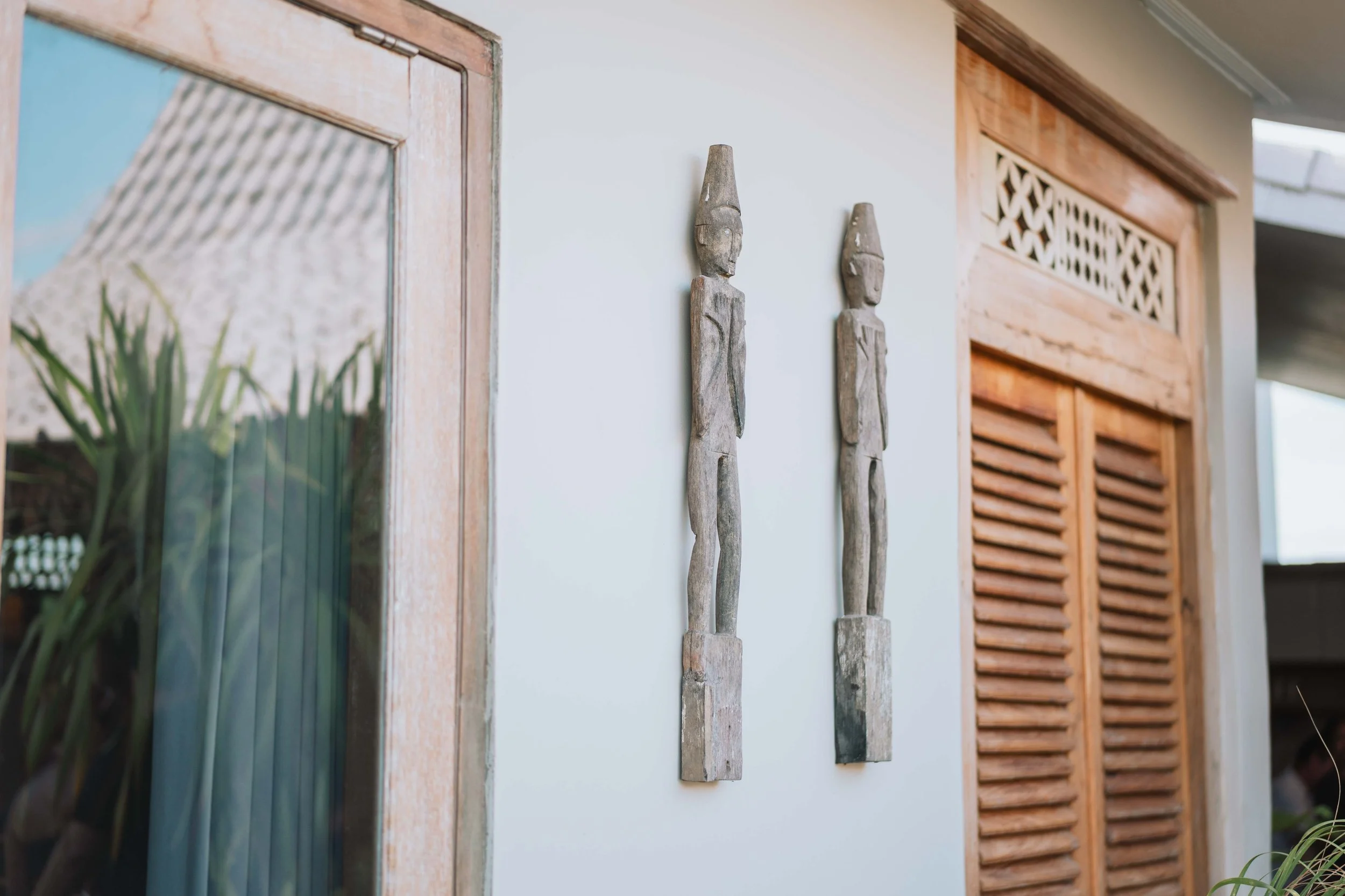 Two wooden tribal sculptures mounted on a white wall in a room with wooden framed windows and doors.