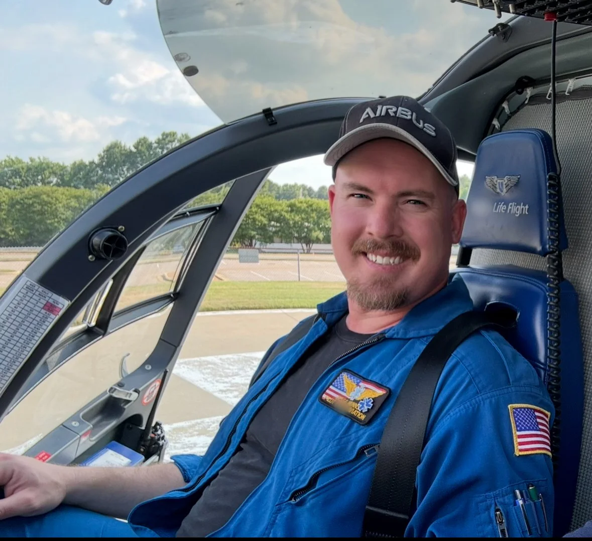 A smiling man in a blue flight suit and cap sitting in a helicopter cockpit.