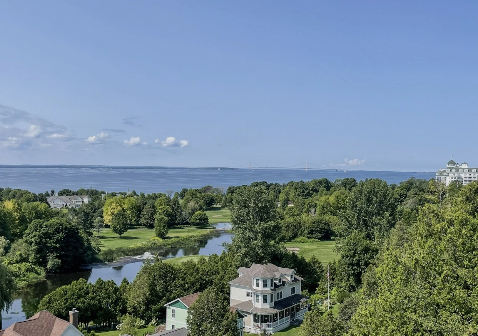 A scenic view of a green landscape with a river, trees, houses, and a distant bridge over a body of water under a bright blue sky with some clouds. High quality drone and aerial photography.
