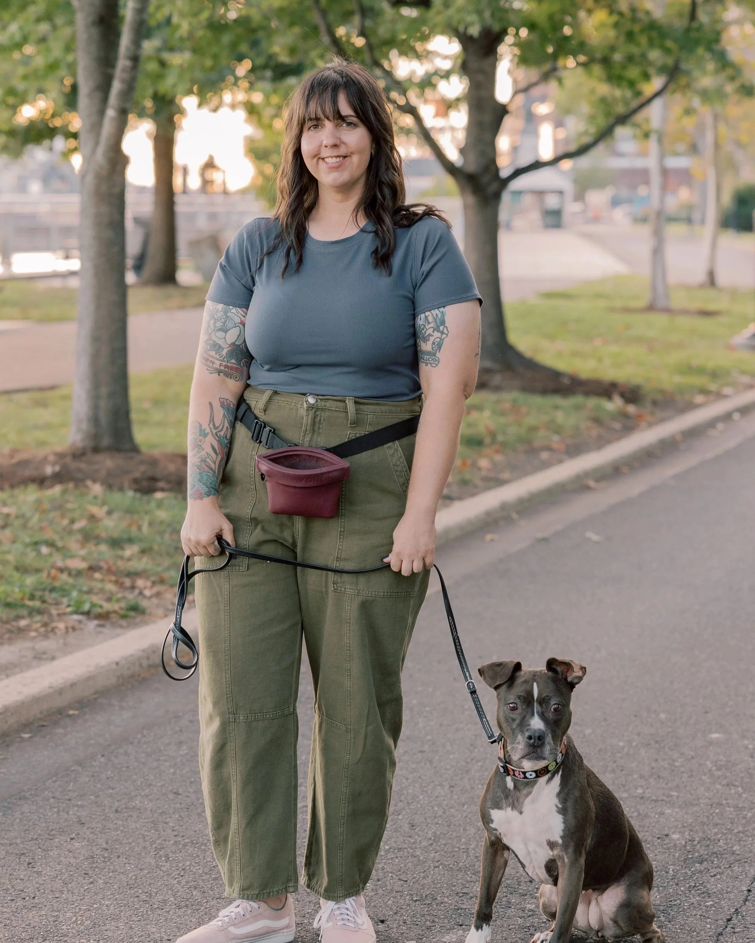 A woman with dark brown hair, wearing a blue T-shirt, olive green pants, and white sneakers, standing on a sidewalk with a leash in her hand, smiling at the camera, as her black and white pit bull mix dog sits beside her on a leash. The background shows trees with green leaves and a park area, during the daytime.