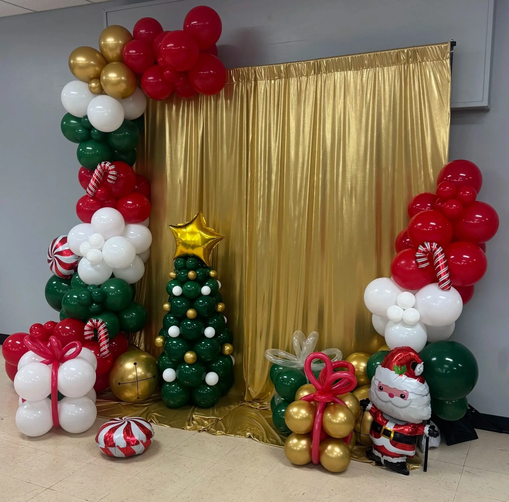Decorative Christmas balloon display with a gold curtain backdrop. Contains balloons in red, white, green, and gold, including a small balloon Christmas tree, a Santa Claus balloon figure, and holiday-themed balloon arrangements.