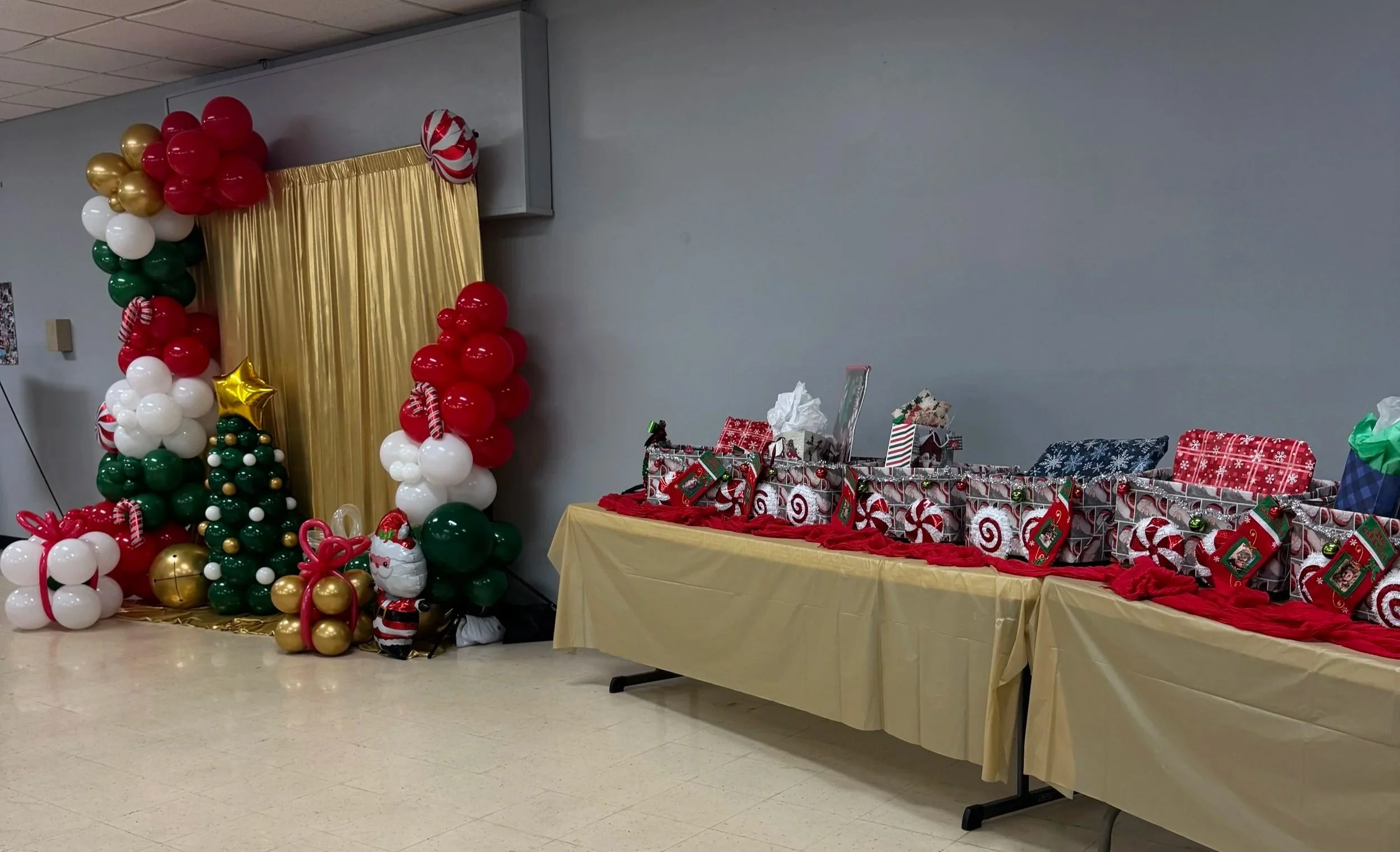 Christmas balloon decorations with a gold curtain backdrop, decorated Christmas tree, and a table with wrapped holiday gifts for gift exchange.