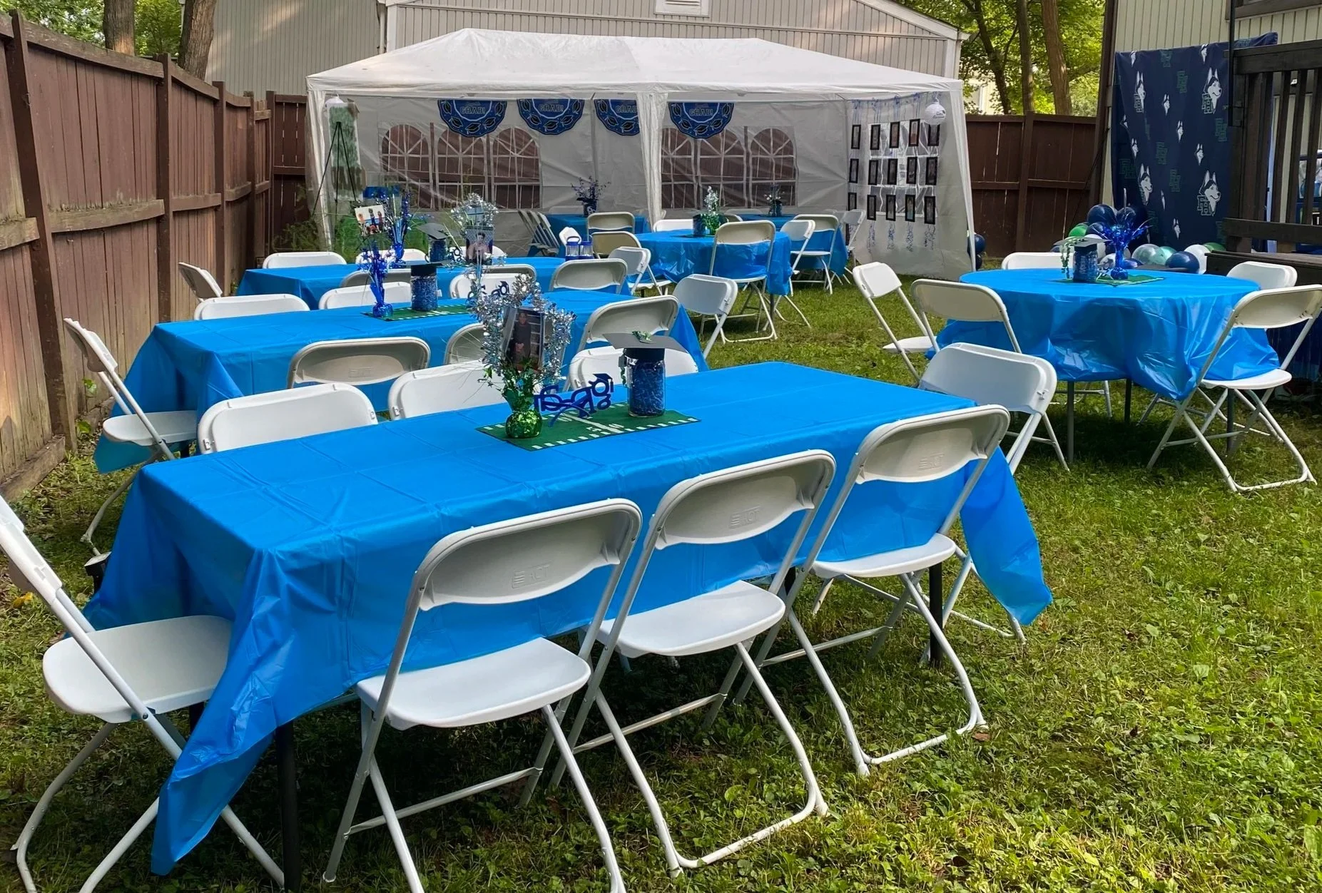 Outdoor backyard graduation table setup with white tent.  Tables are decorated centerpieces on blue tablecloths, white folding chairs, and decorations for a graduation celebration.