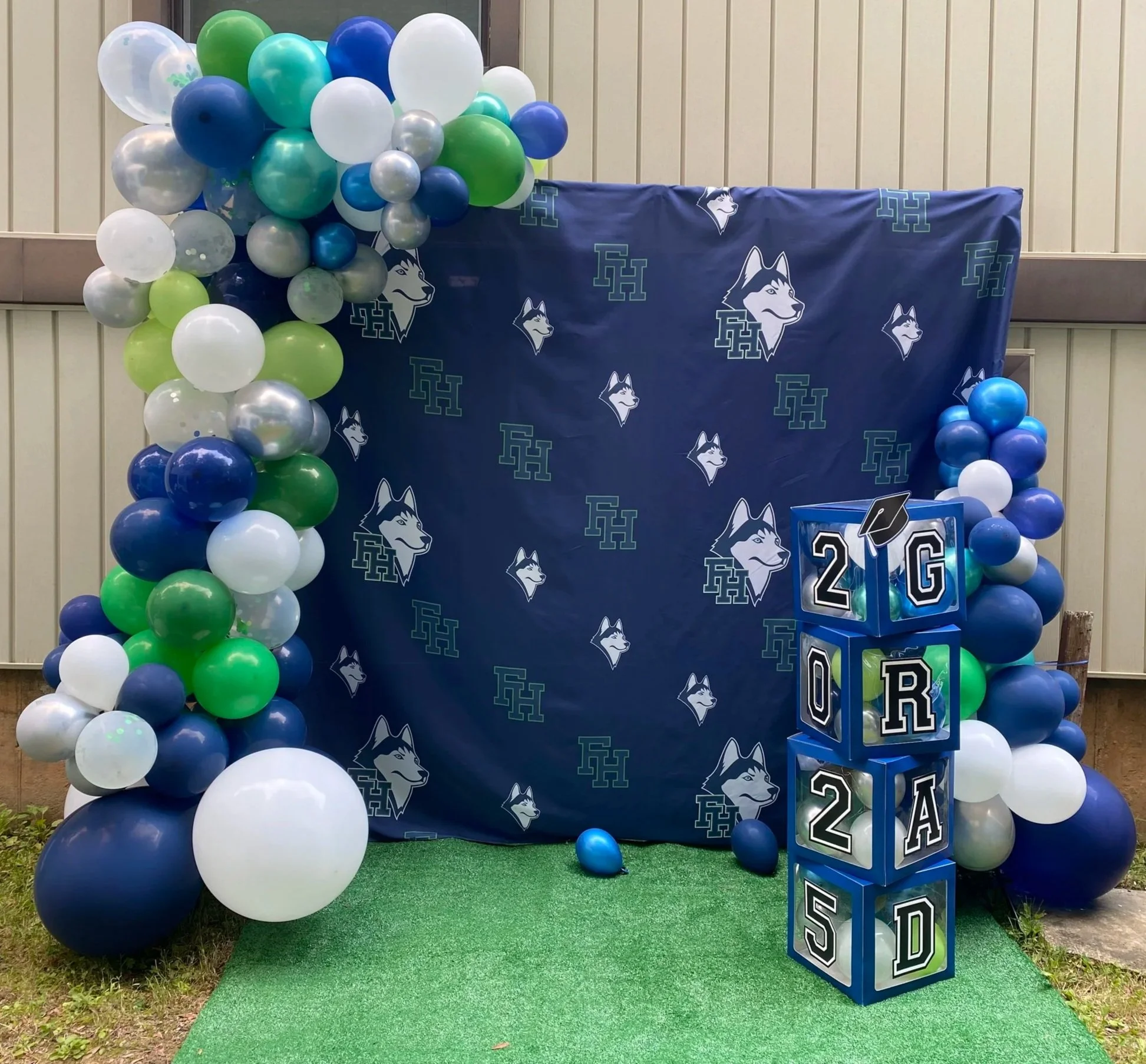 Graduation-themed backdrop with balloons in blue, green, white, gray, and silver, arranged in a column on each side of the school backdrop, with branded boxes spelling '2025' and a graduation cap decoration, all on a green artificial turf.