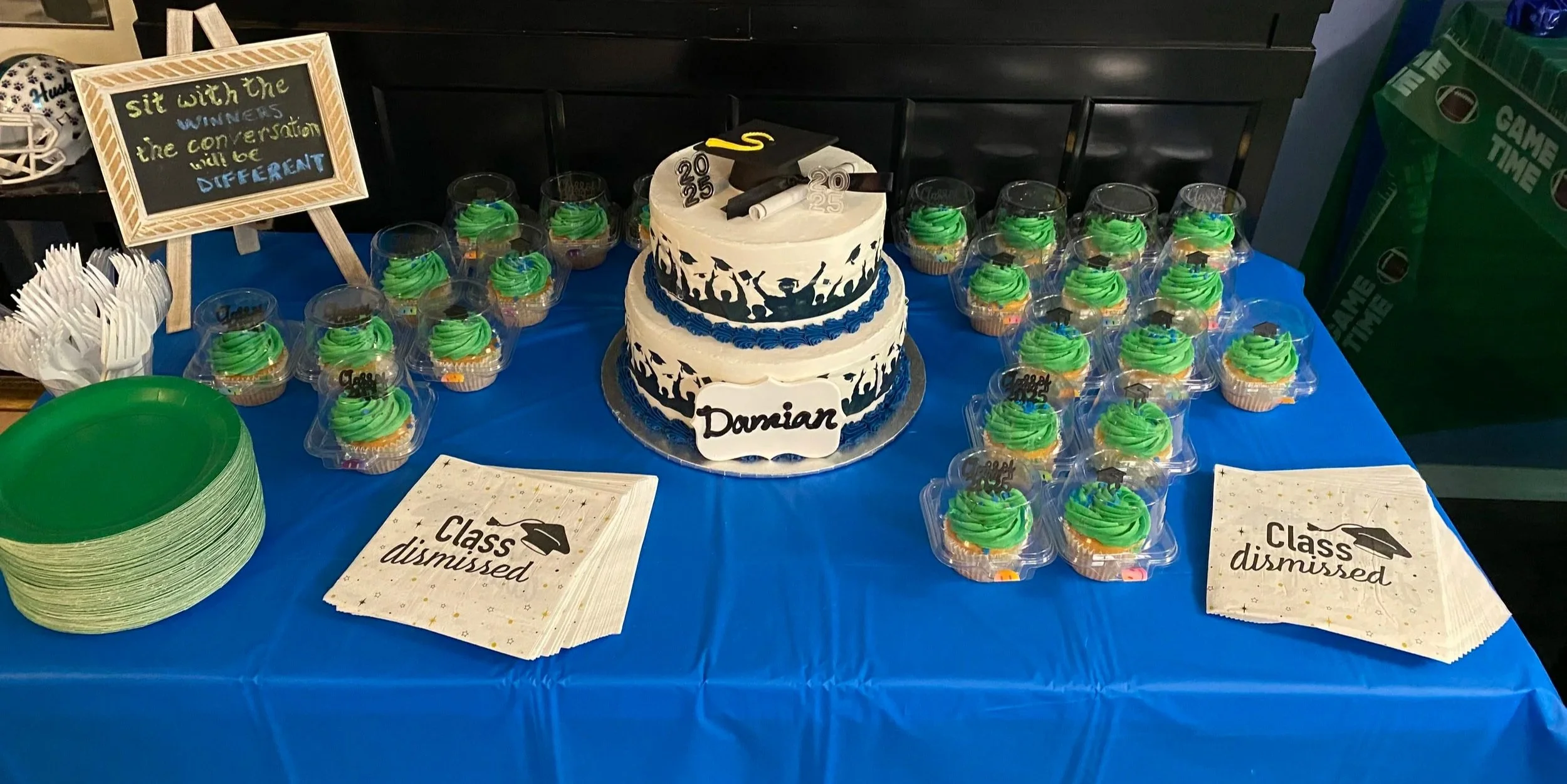 Graduation-themed dessert table with a two-tiered cake decorated with graduation caps, diplomas, and a graduate silhouette, surrounded by cupcakes with green frosting and graduation toppers, and 'Class dismissed' napkins, on a blue tablecloth.