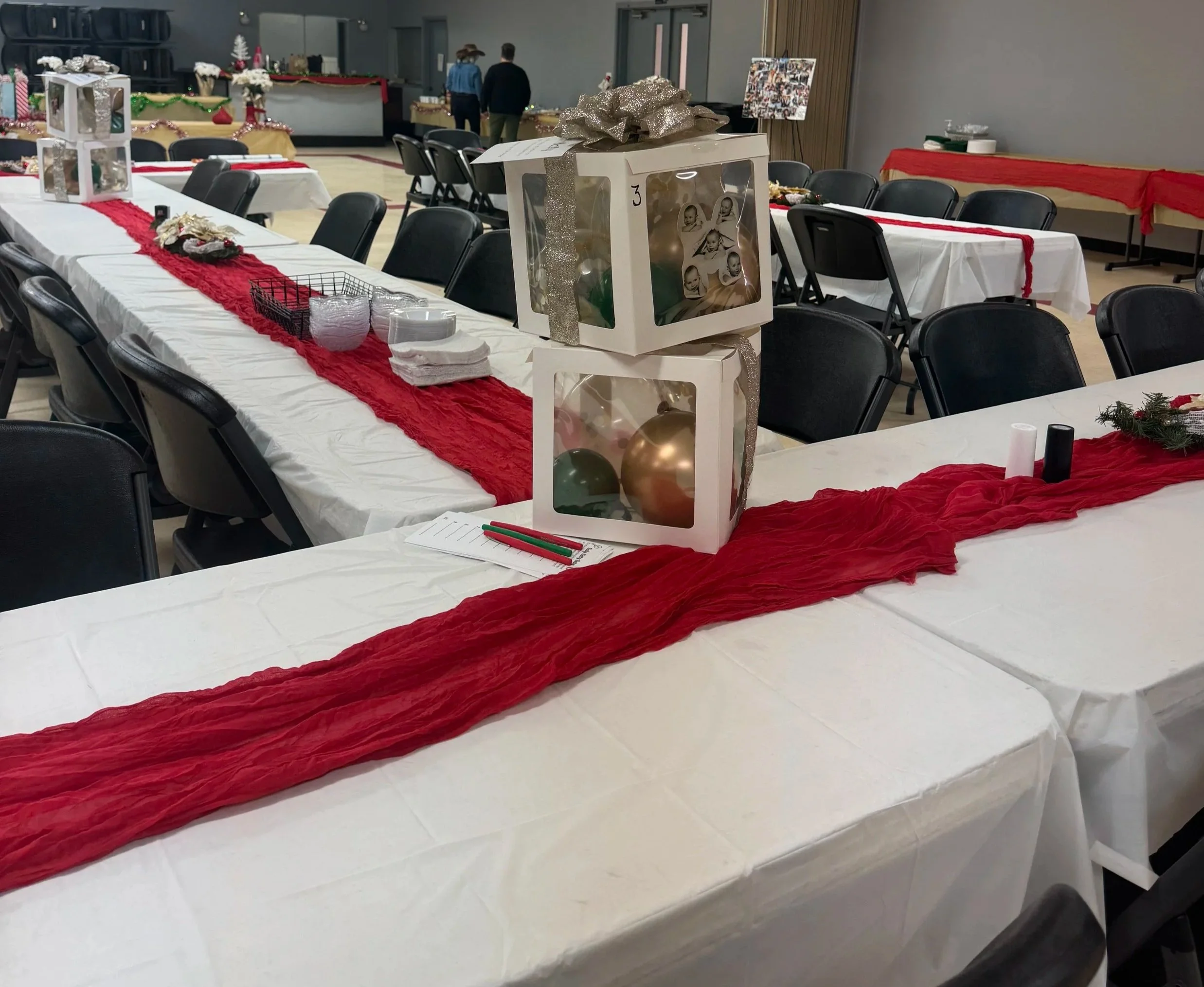 Decorated dining hall with long tables covered in white tablecloths, red table runners, and holiday decorations including gift boxes and ornaments, likely set for a Christmas celebration.