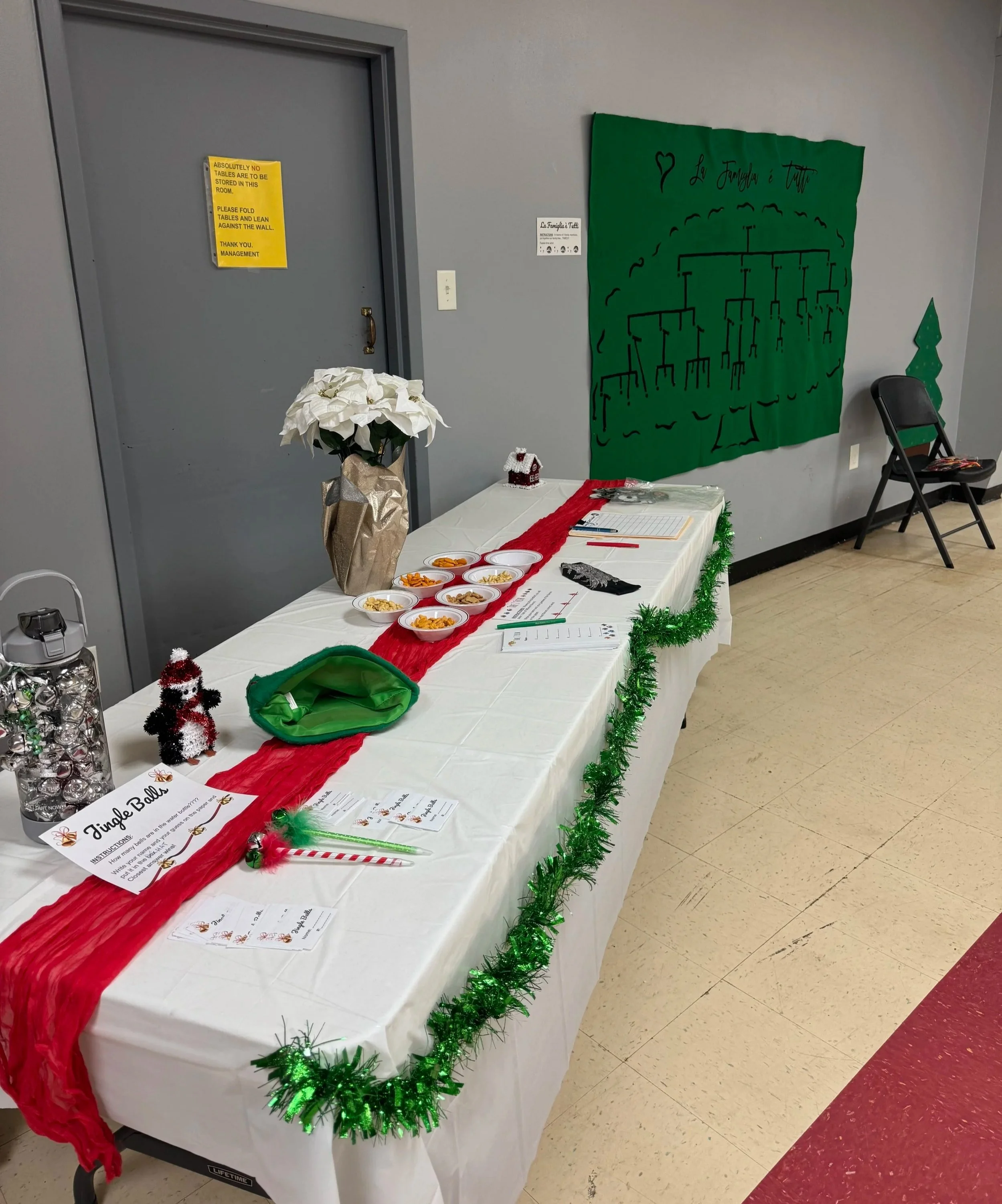 A decorated table with a white tablecloth, red runner, and green tinsel garland. Small bowls with snacks, holiday decorations, and sign-in sheets are on the table. Behind the table is a green Christmas themed mural and a black chair. There are holida