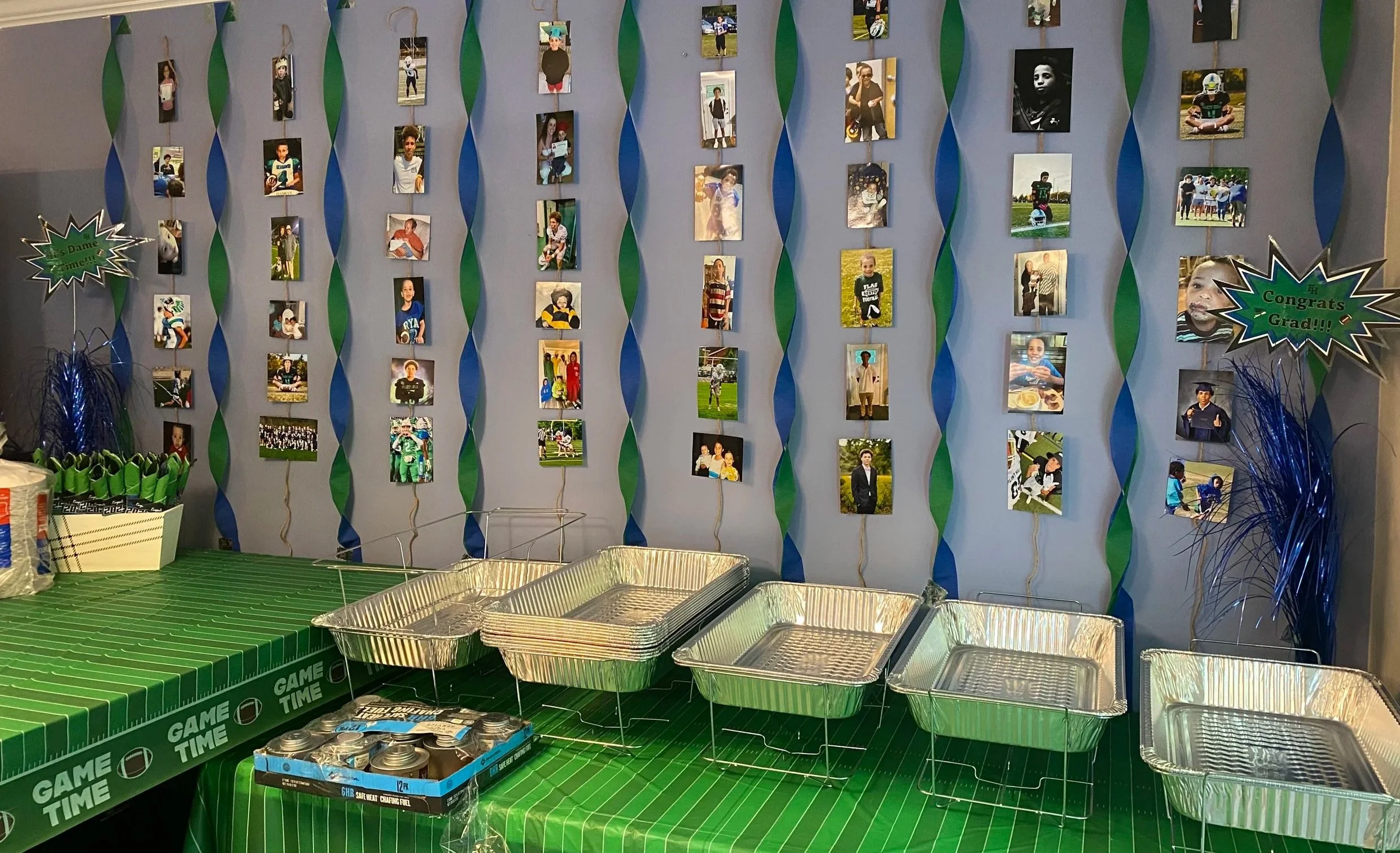Decorated wall with hanging photos of children, blue and green streamers, and congratulatory signs; table with empty aluminum trays, a green tablecloth with sports-themed print, and party supplies.