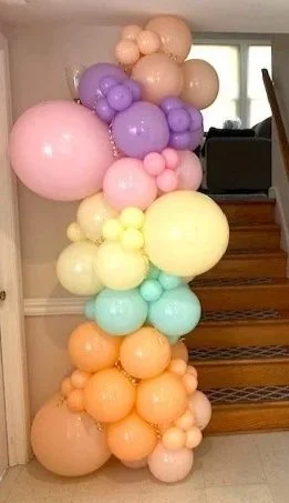 A column of pastel-colored balloons arranged along a staircase wall, featuring shades of pink, purple, yellow, mint green, and peach.