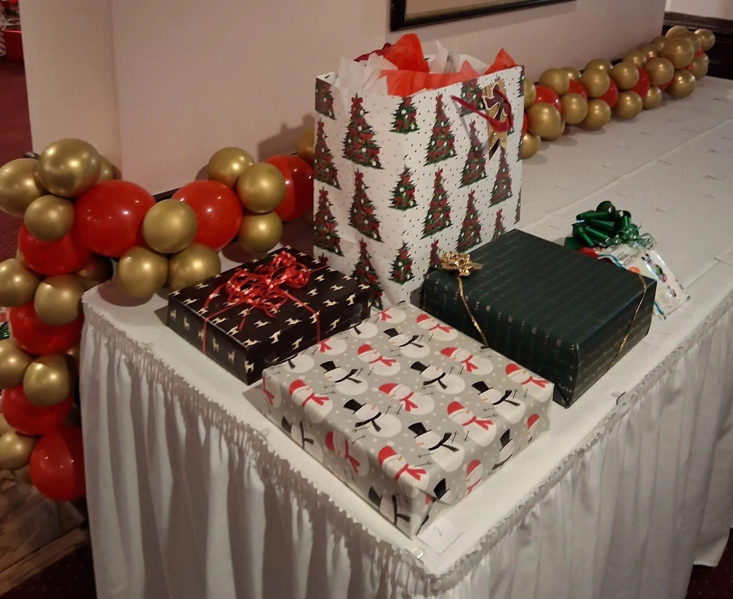 Christmas presents/ gift table on a white tablecloth-covered table, decorated with holiday themed gifts surrounded by a balloon garland with gold and red balloons.