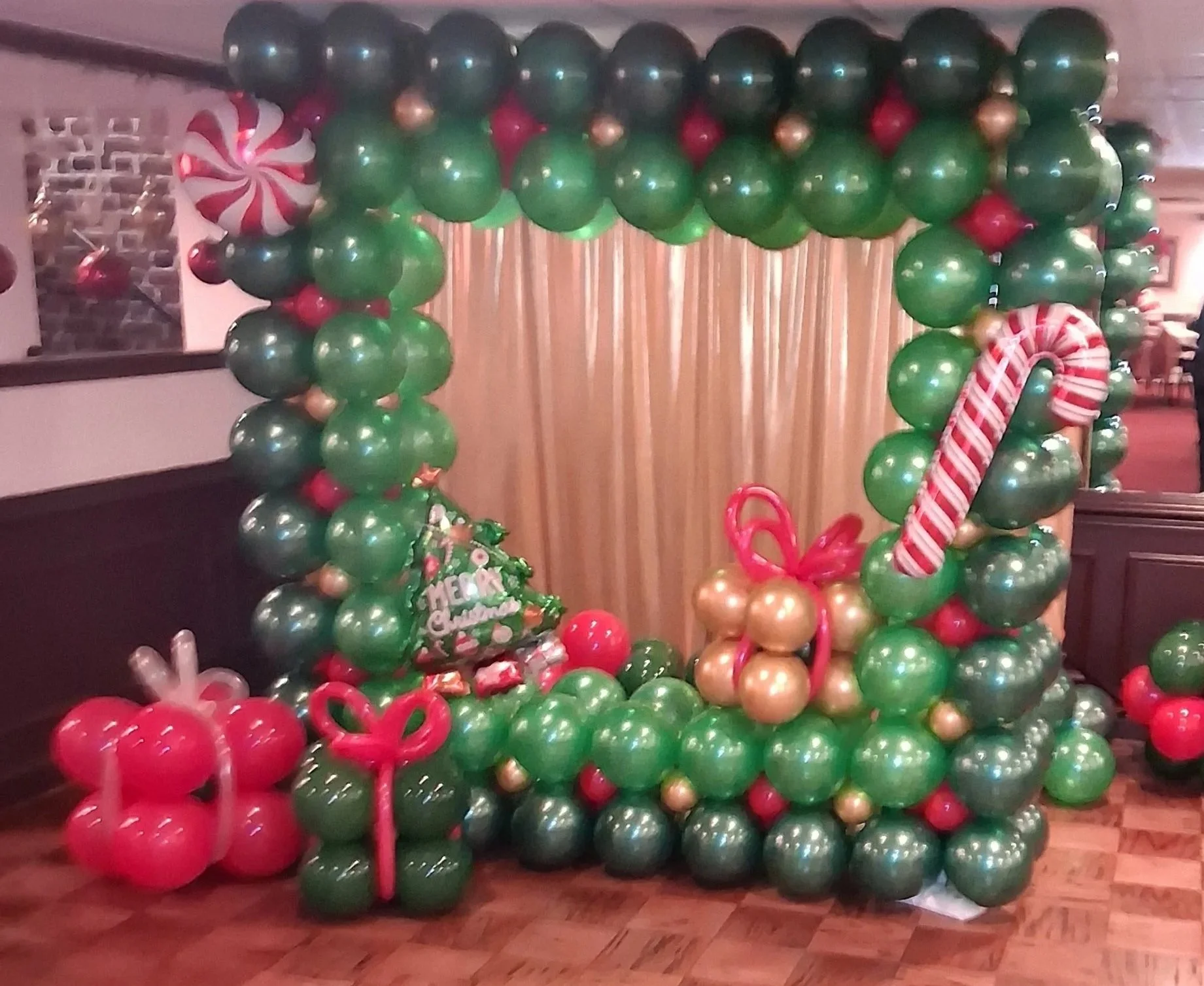Christmas balloon picture frame display with green, red, and gold.  Blowup candy canes, peppermints, and a Christmas tree in a festive setting with balloon presents in front of a gold backdrop.