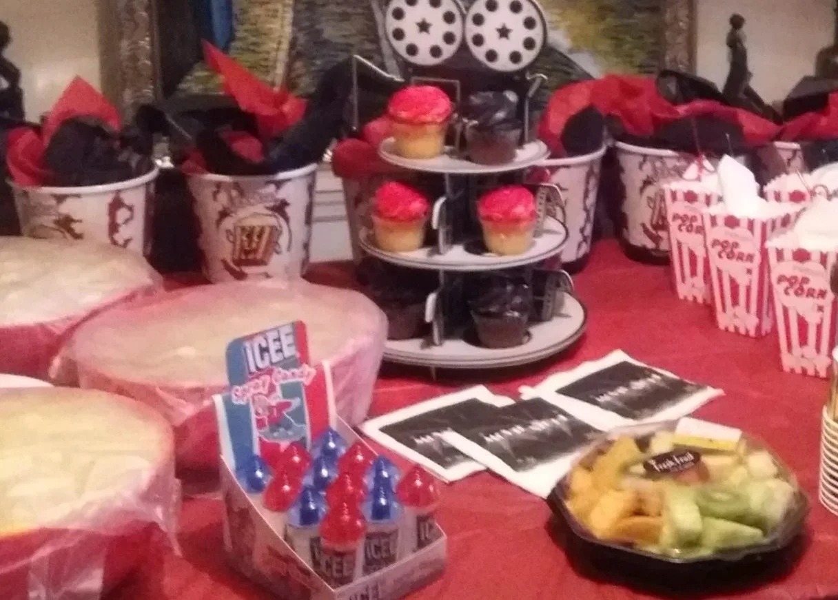 A dessert and snack table decorated for a movie themed birthday celebration, featuring cupcakes with red frosting, popcorn in red and white striped containers, icee slush beverages, and chocolate-covered pretzels for movie night.
