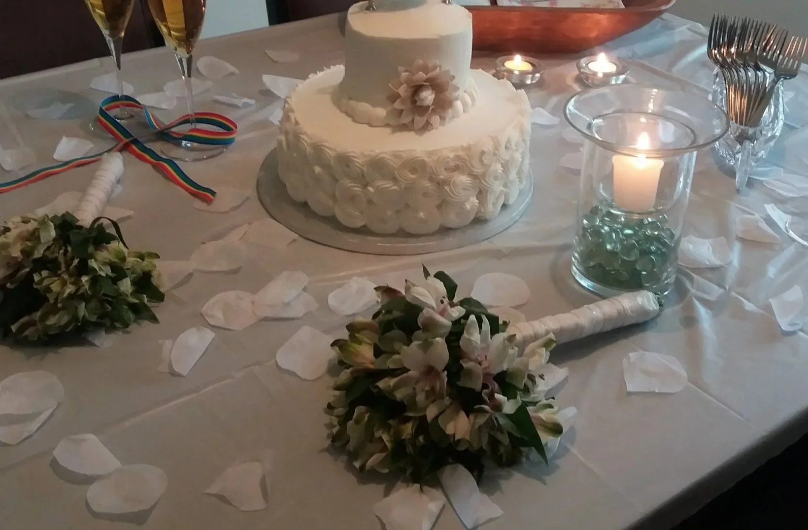 A decorated table featuring a white 2 Tier Wedding cake surrounded by white petals and 2 floral bouquets, candles, glasses, and cutlery, suggesting a LGBQT reception.