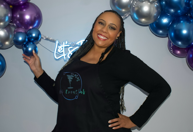 A woman smiling and posing in front of a balloon decoration and neon sign, wearing a black apron with a 'BS Eventful' logo and text, in an indoor setting.