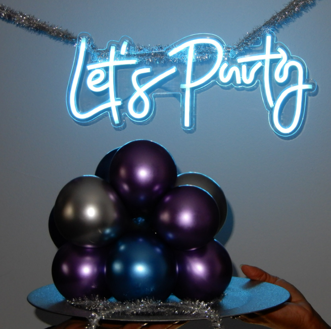 Decorative party display with a neon sign saying 'Let's Party', metallic balloons in purple, black, and blue, and tinsel.