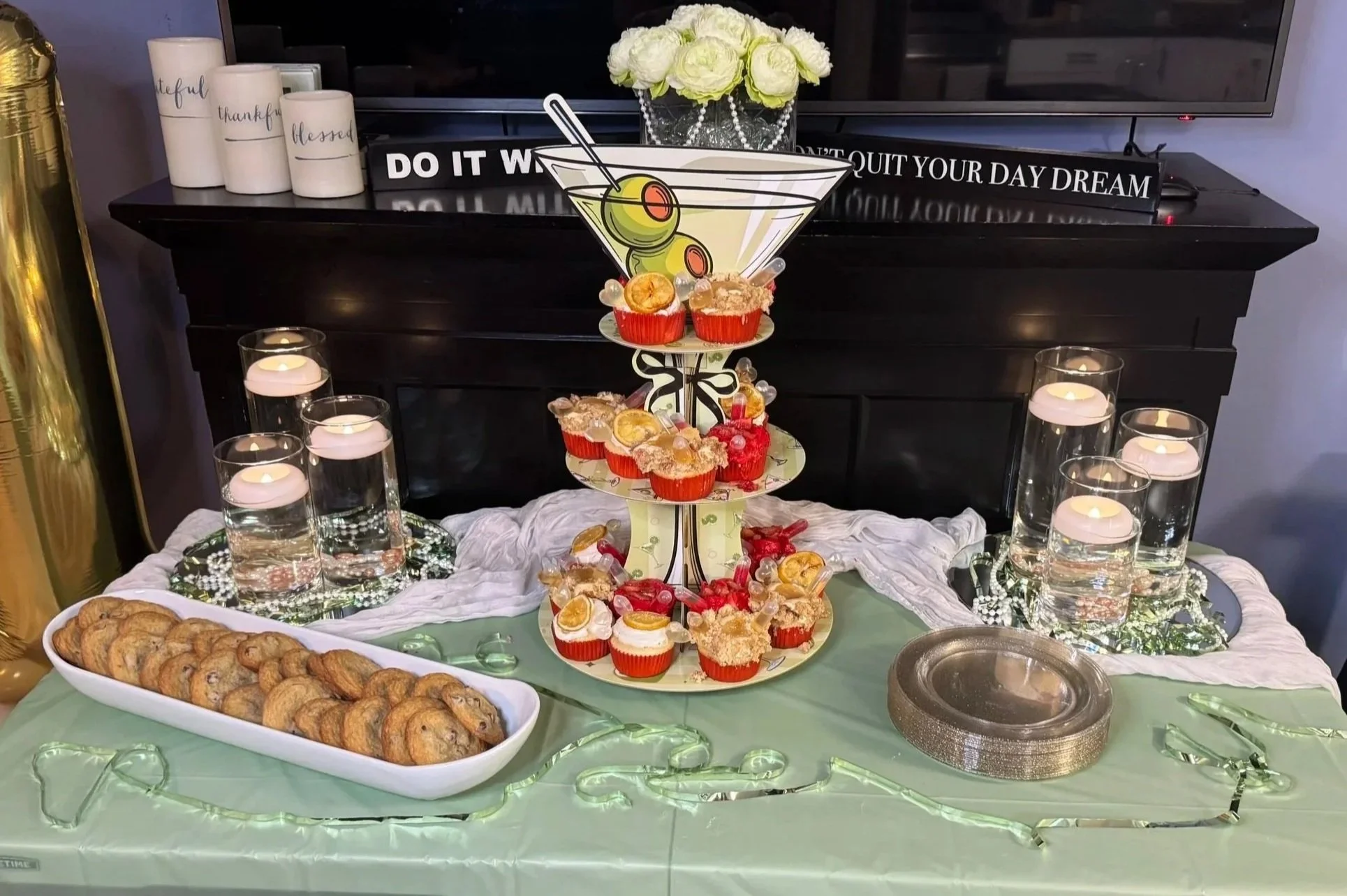 A dessert table with cookies, cupcakes, and a three-tiered stand with candies, decorated with a cocktail-themed sign. Candles and decorative lights are also on the table.