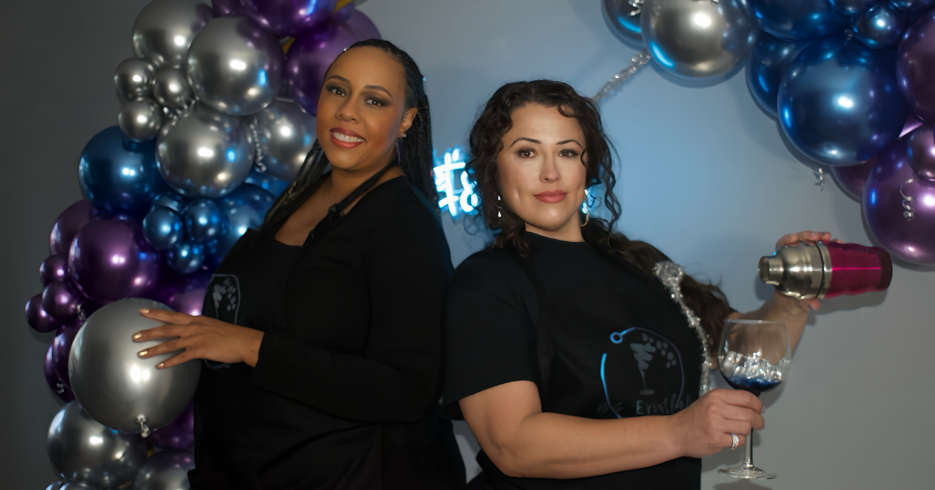 Two women stand in front of a colorful balloon arch, one holding a silver balloon and the other pouring a beverage from a pink shaker into a glass.