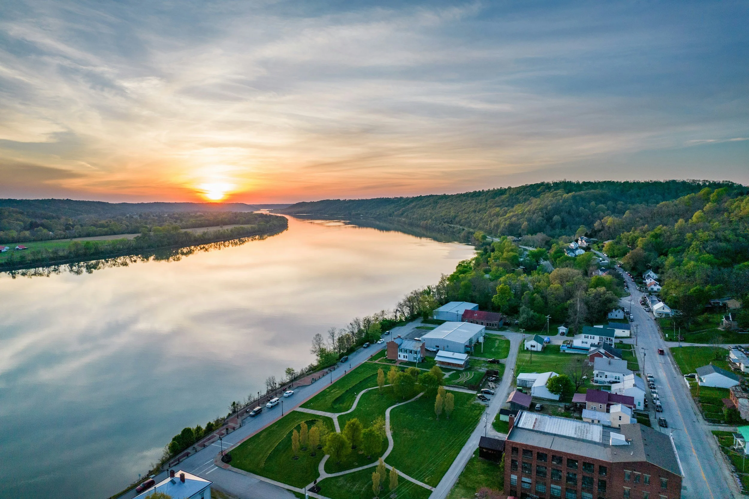 Aerial view of a river at sunset with a small town along the riverbank, surrounded by green hills and trees.