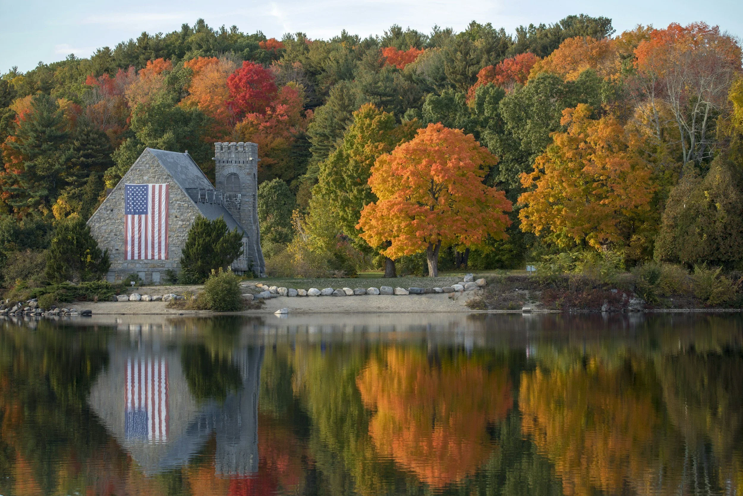 A stone church with American flag decoration stands by a lake, surrounded by colorful fall foliage in red, orange, yellow, and green, with trees reflected in the water.