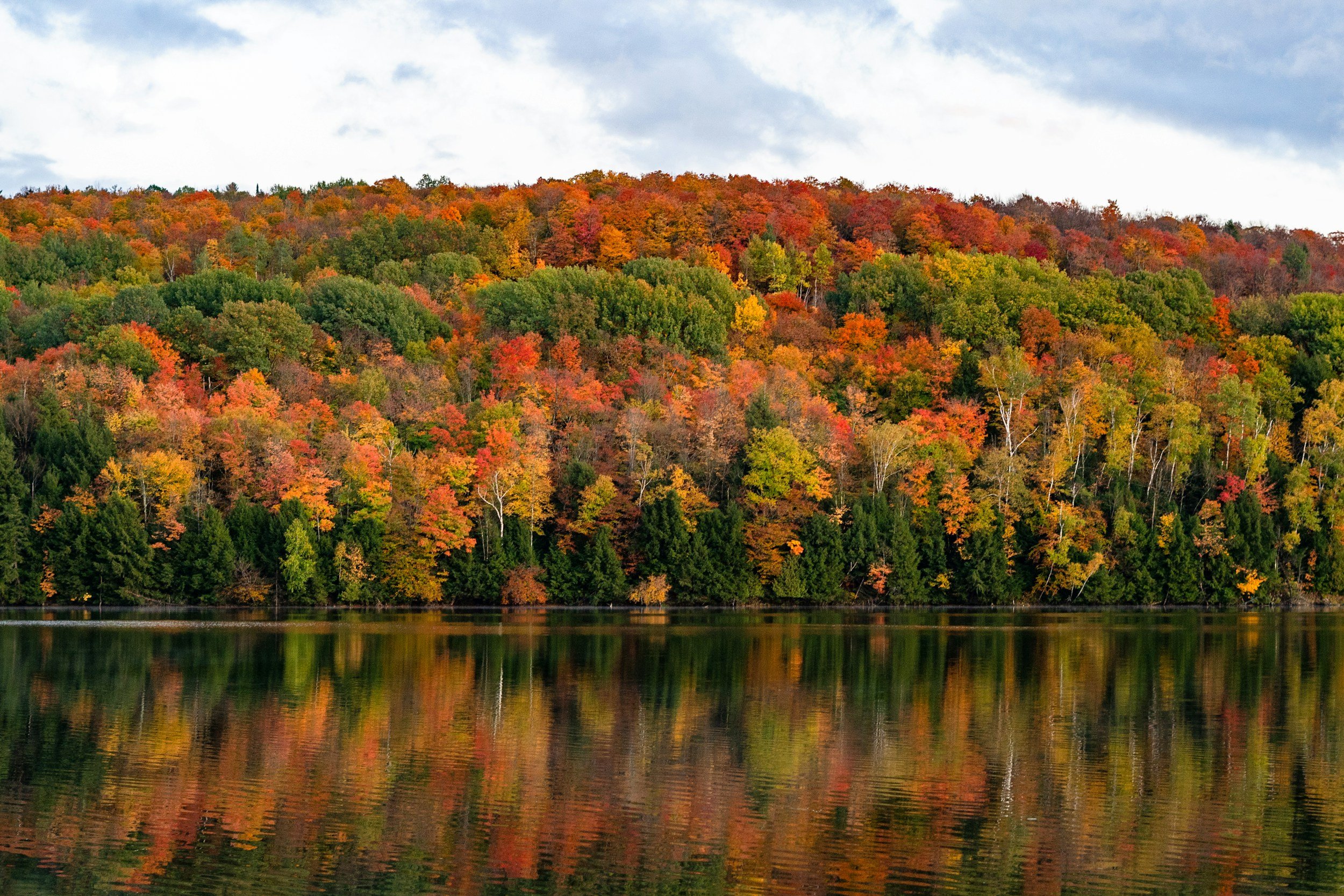 Colorful fall foliage on a hillside reflected in a lake, with a partly cloudy sky.