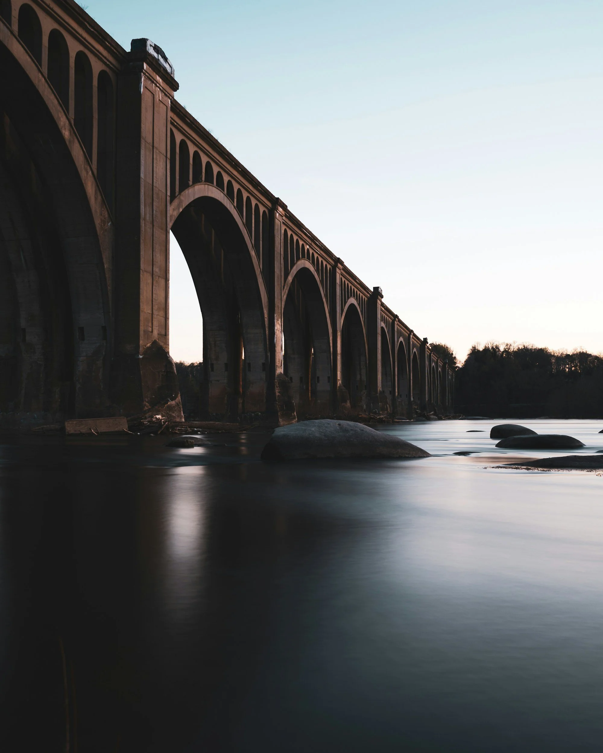 A large brick arch bridge crossing over a calm river at dusk with rocks in the water and trees in the background.