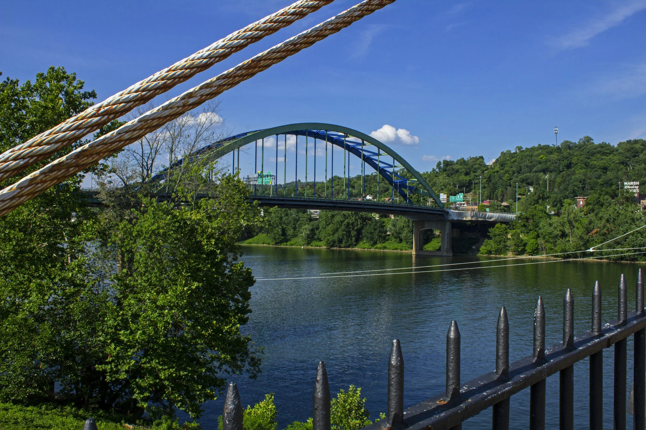 View of a bridge over a river, with greenery and trees on both sides, and a bright blue sky with a few clouds.