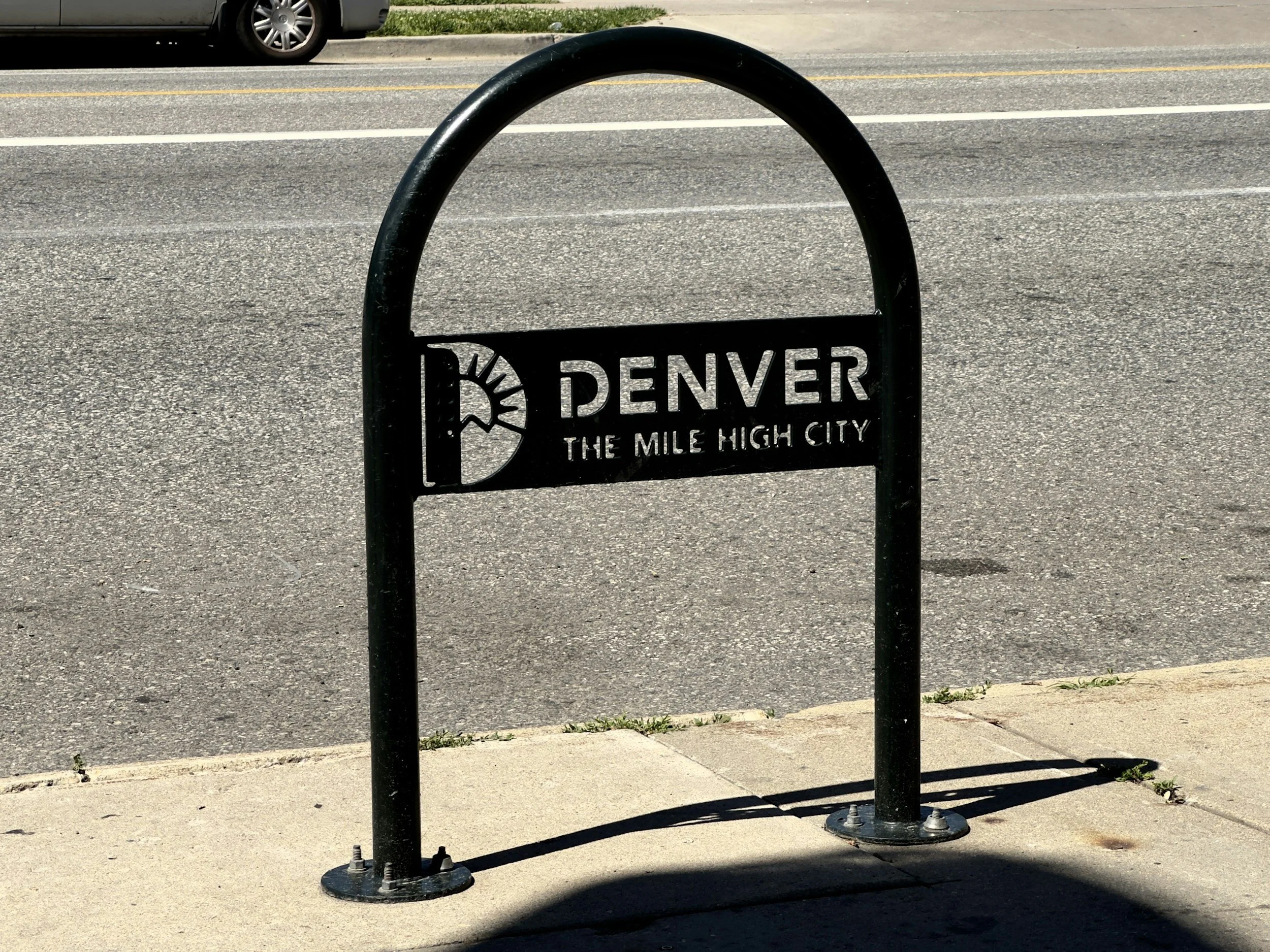 Black bike rack on a sidewalk with a sign that reads "Denver The Mile High City" and the Denver logo, with a road and a vehicle in the background.