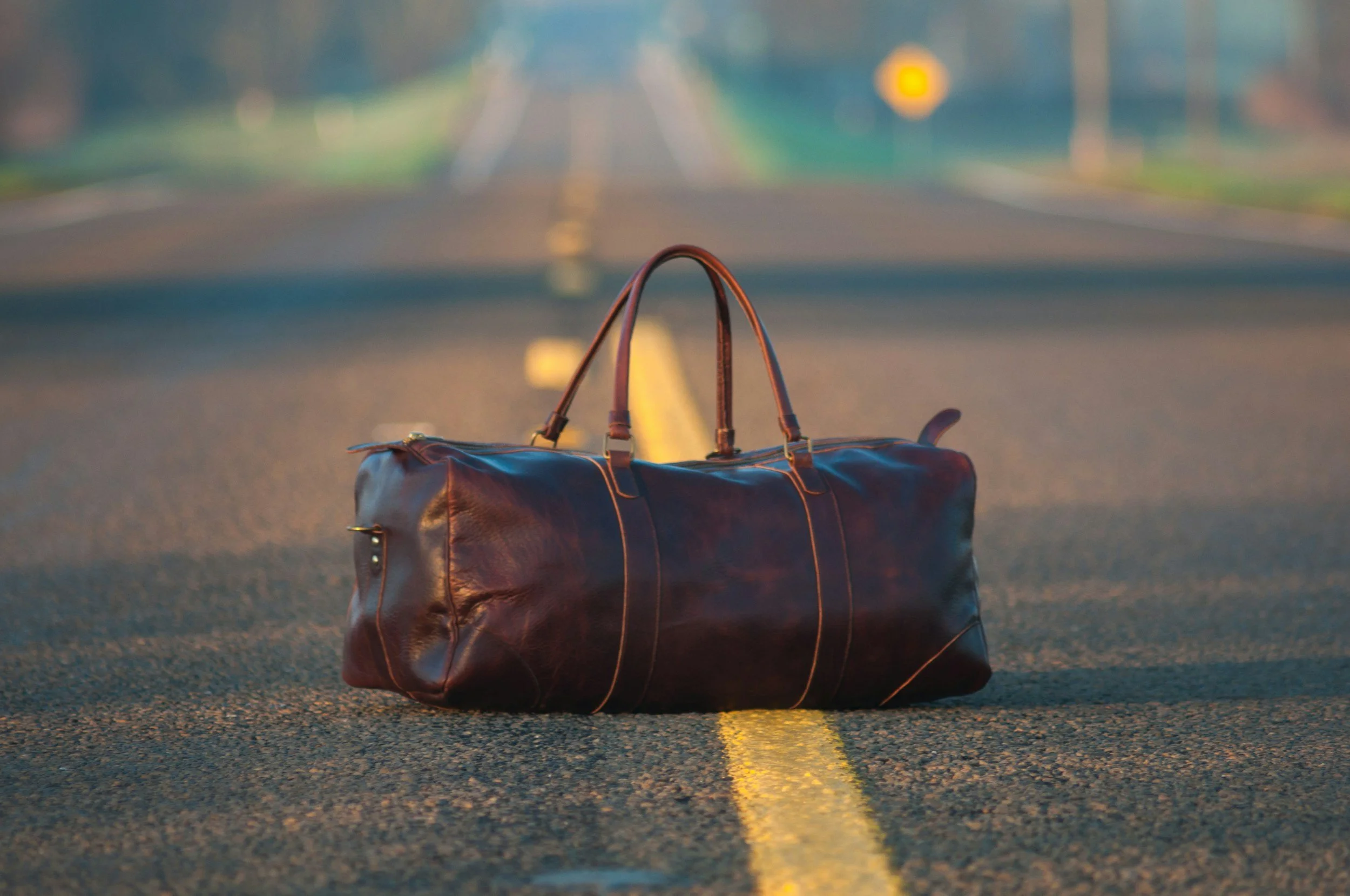 Brown leather duffel bag on a deserted road with yellow center lines, slightly blurred background of trees and distant horizon.