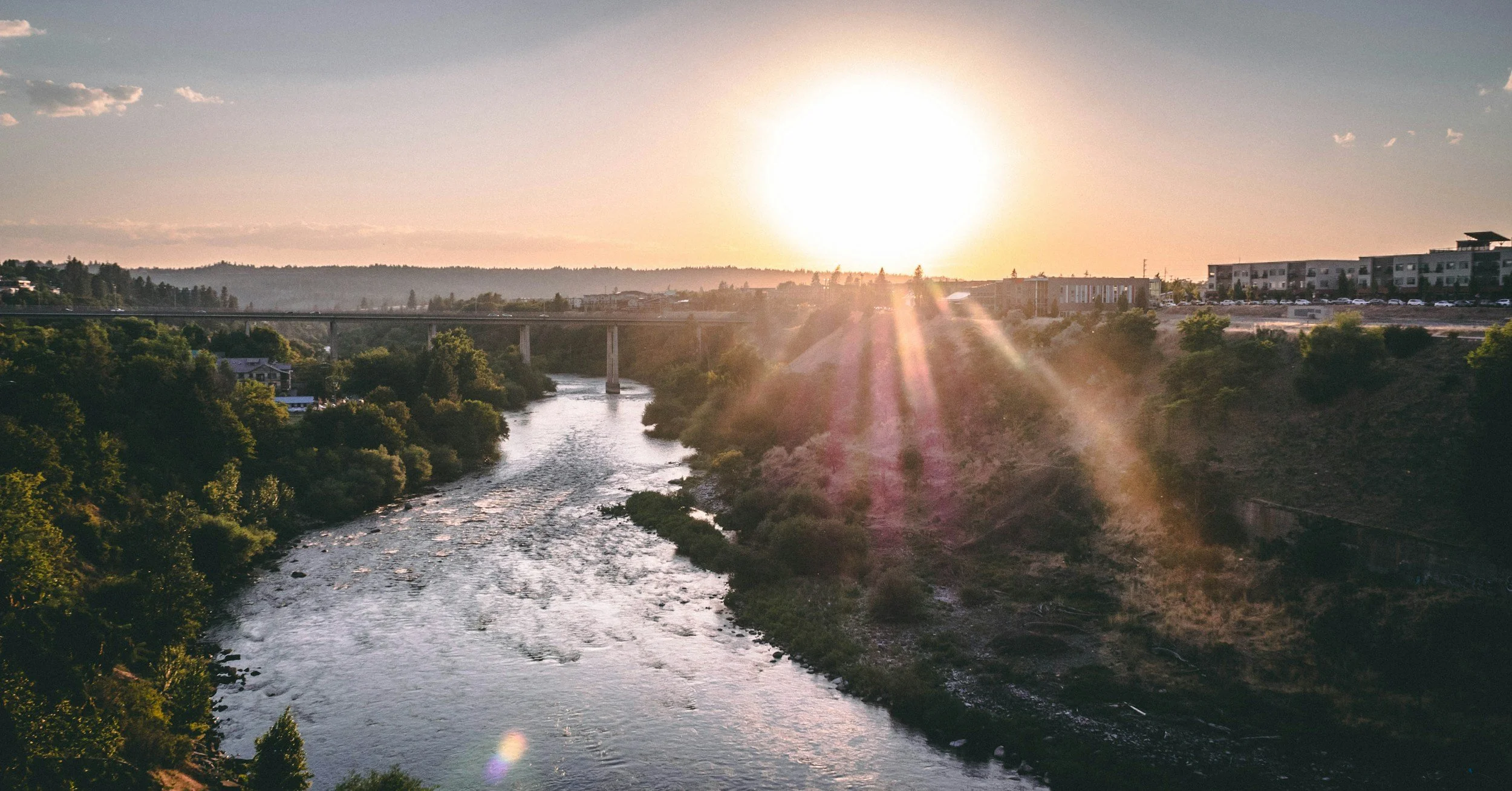 A river flowing through a valley with lush green trees on both sides, with a bridge crossing over the river; the sun is setting in the background, casting a warm glow and creating lens flare effects.