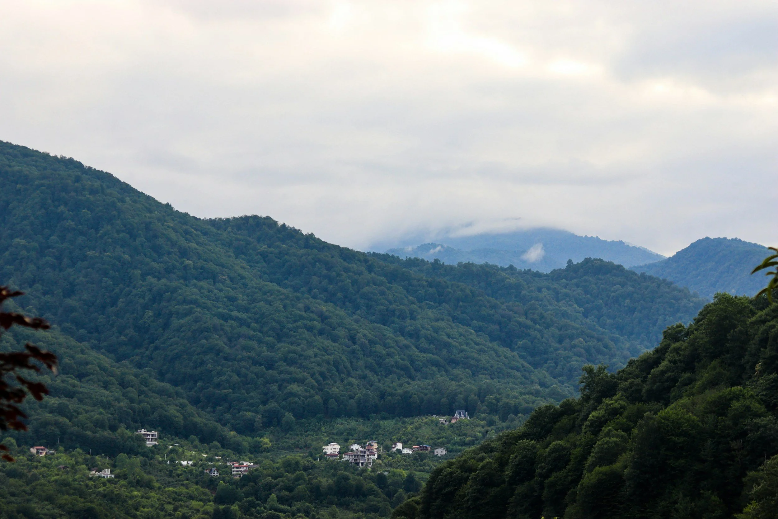 Green forested mountains with a few houses visible at the base, cloudy sky with mist on the mountain peaks.