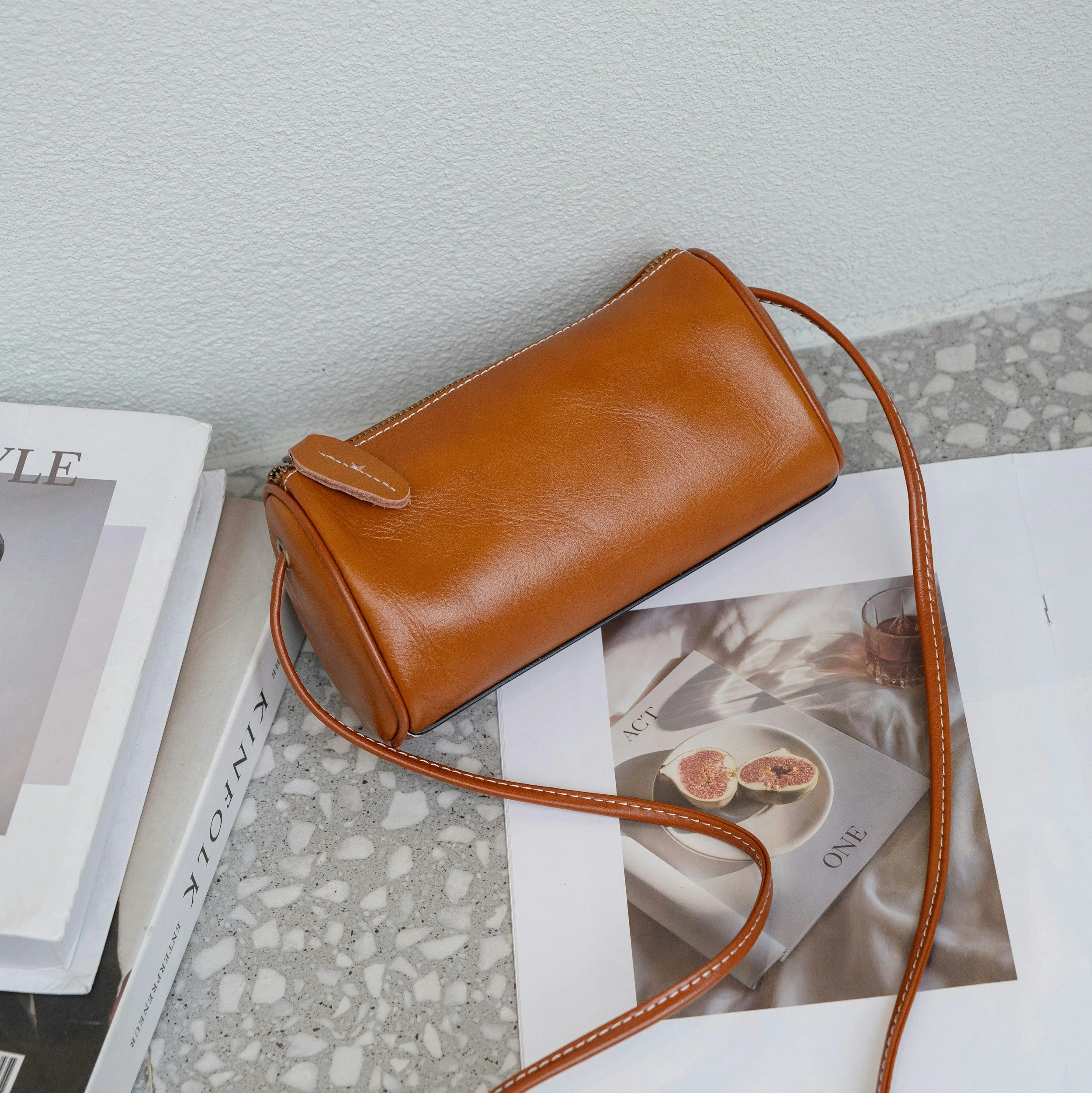 A tan leather handbag resting on open magazines on a speckled gray surface against a light textured wall.