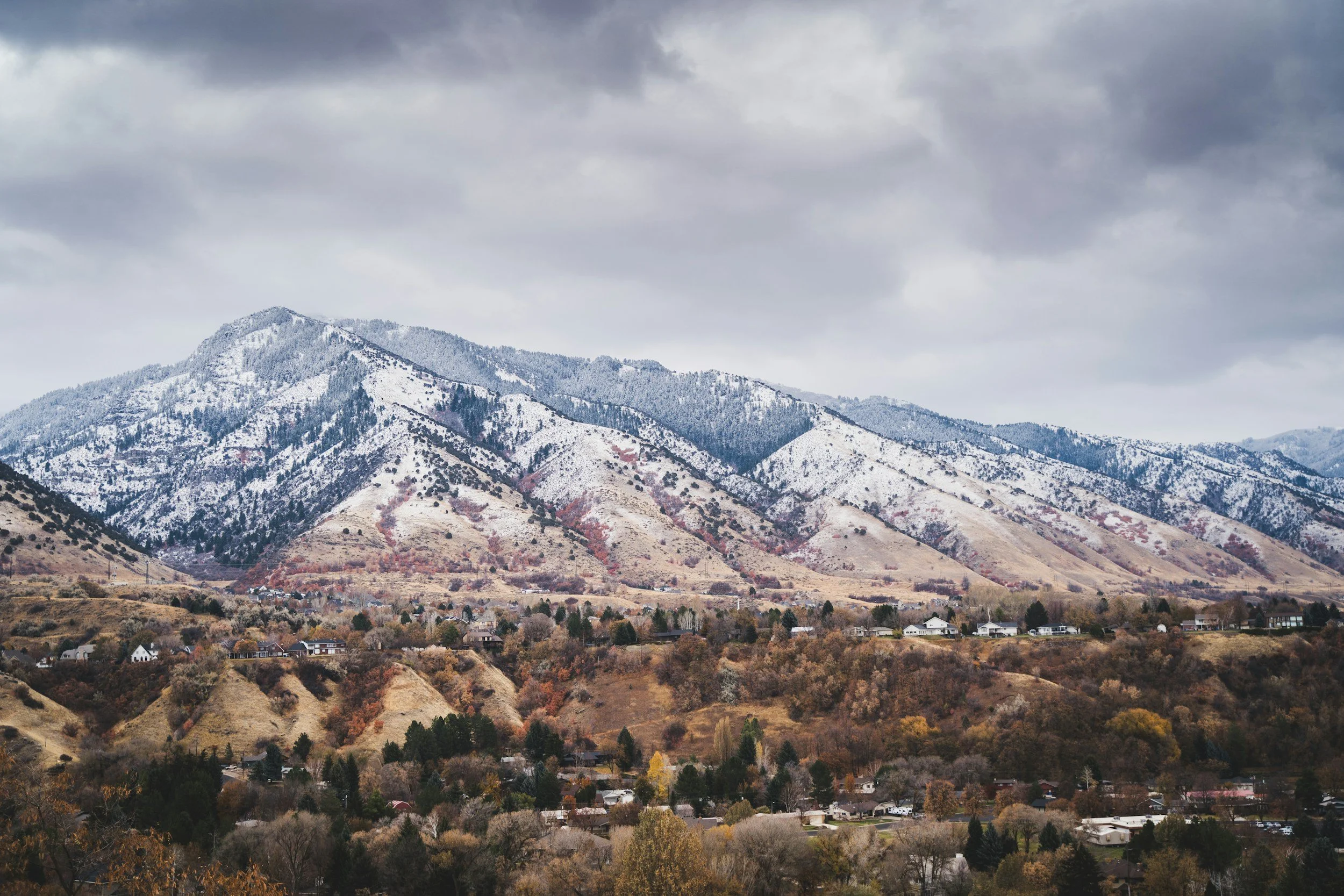 Mountains with snow at the peaks and a small town in the foreground under overcast sky.