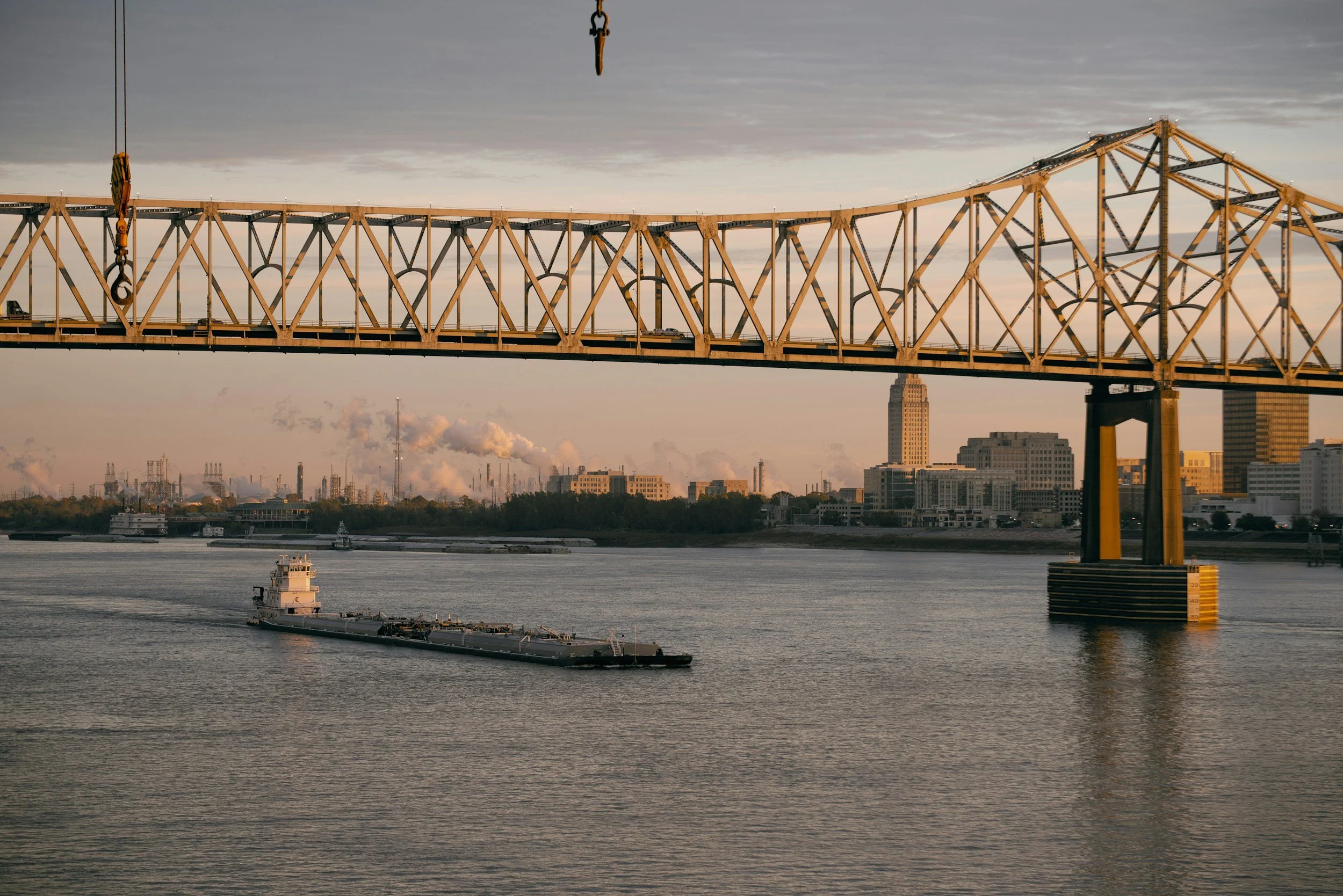 A large crane is suspended above a river with a barge passing underneath. In the background, there are buildings and industrial structures, with some smoke or steam rising into the sky.