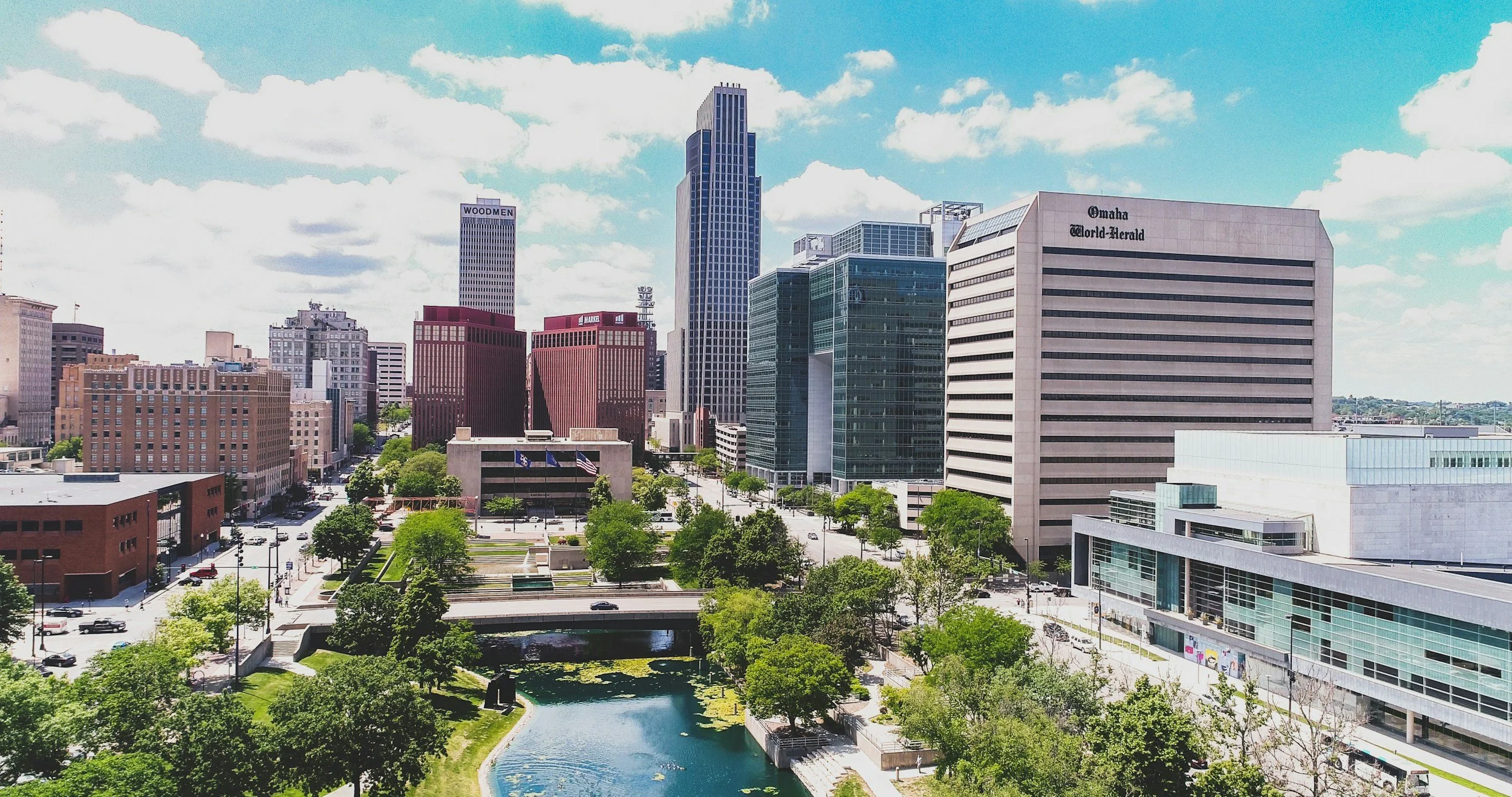 Skyline of Omaha, Nebraska with modern high-rise buildings, a park with a pond, and traffic on nearby streets under a partly cloudy sky.