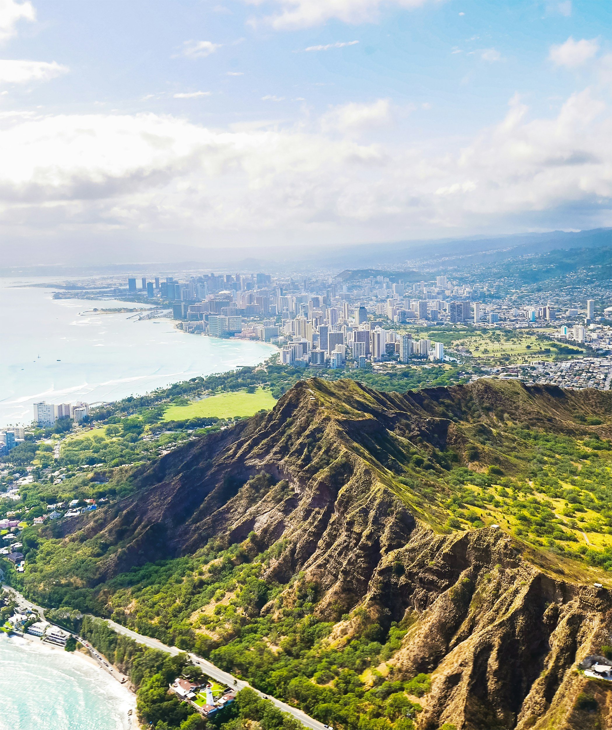 Aerial view of Diamond Head crater with a city skyline and coastline in the background, lush green vegetation in the foreground.