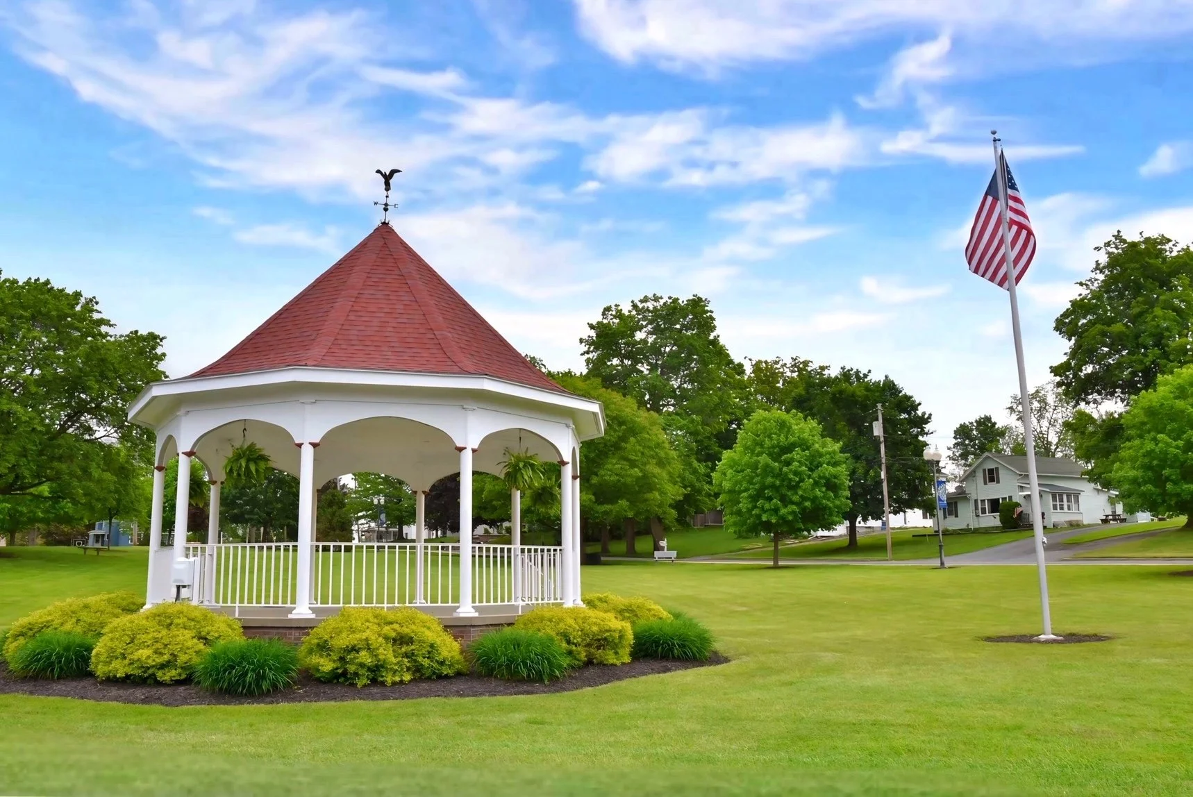 A park scene with a white gazebo, American flag, green trees, and a grassy lawn under a blue sky with clouds.
