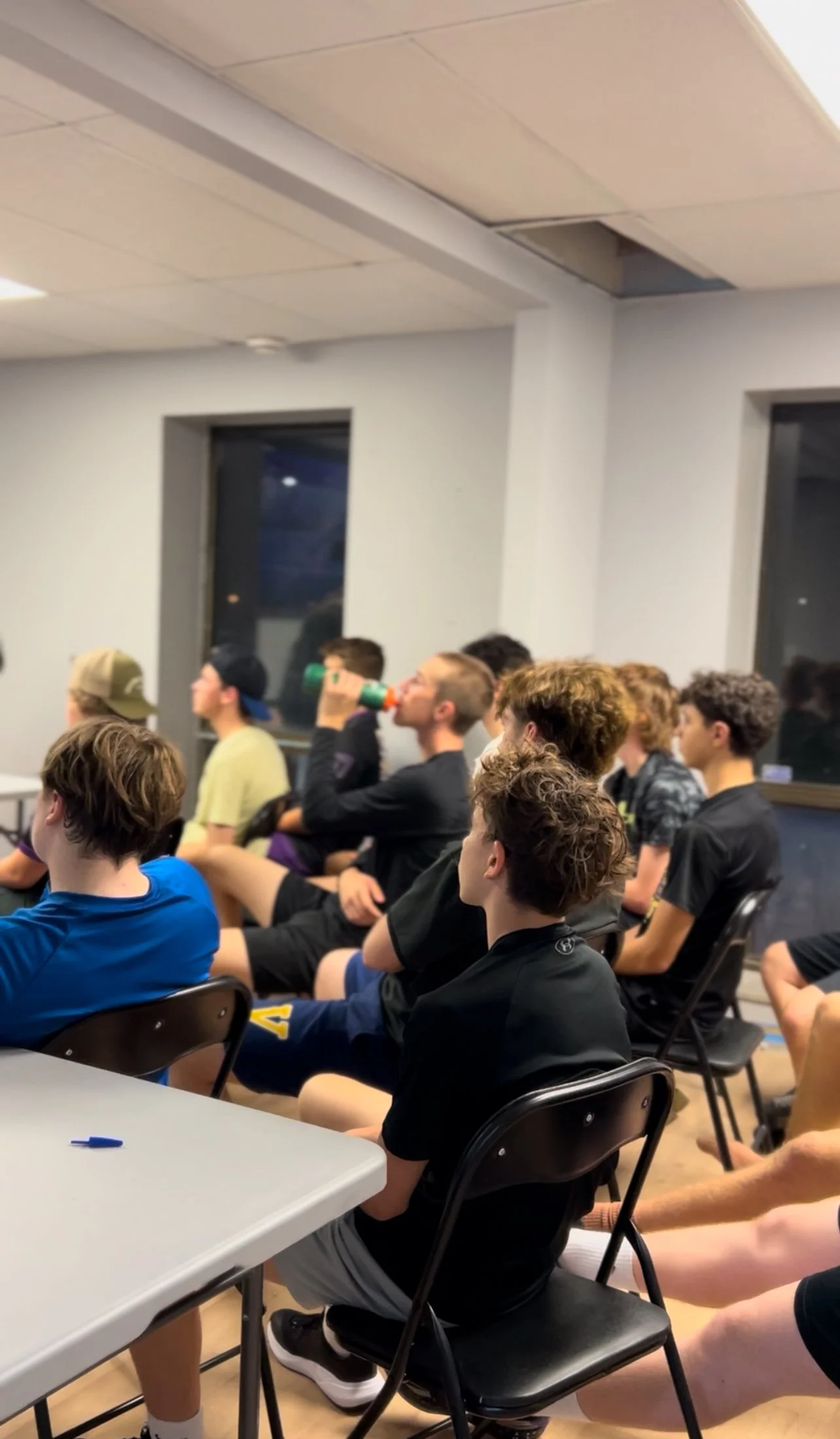 A group of boys seated in a classroom, some watching something off-camera, one drinking from a water bottle. The classroom has gray walls, windows, and a section of the ceiling damaged or open.