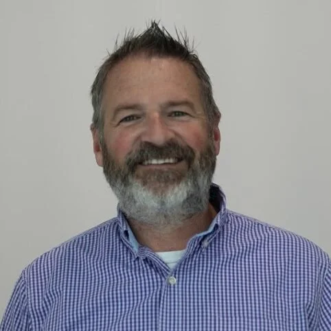 A smiling man with a beard and short, spiky hair, wearing a blue checkered shirt, standing against a plain light background.