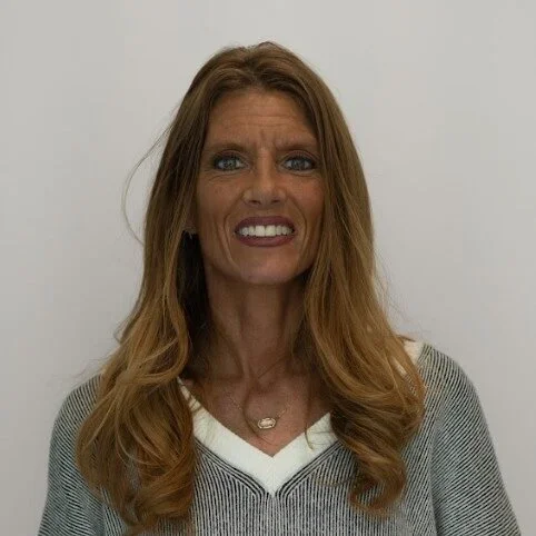 A woman with long, wavy, light brown hair, smiling in front of a plain white background, wearing a gray and white striped sweater and a gold necklace.