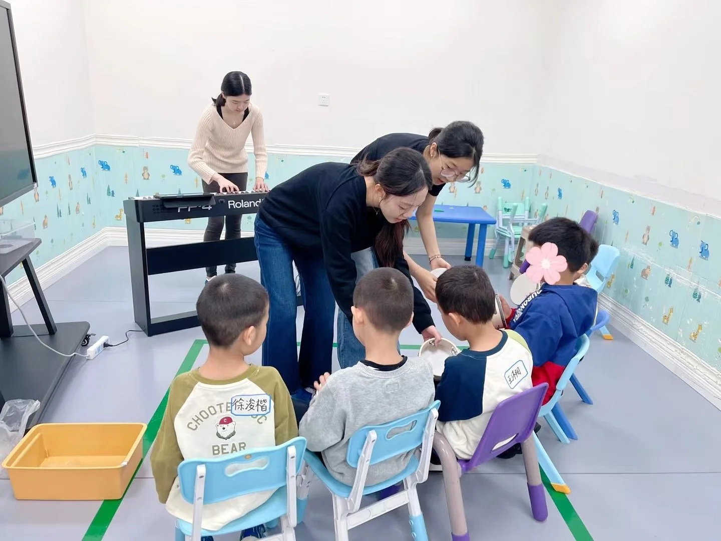 Children sitting at a table participating in a classroom activity with teachers assisting, in a decorated room with colorful chairs and wallpaper.