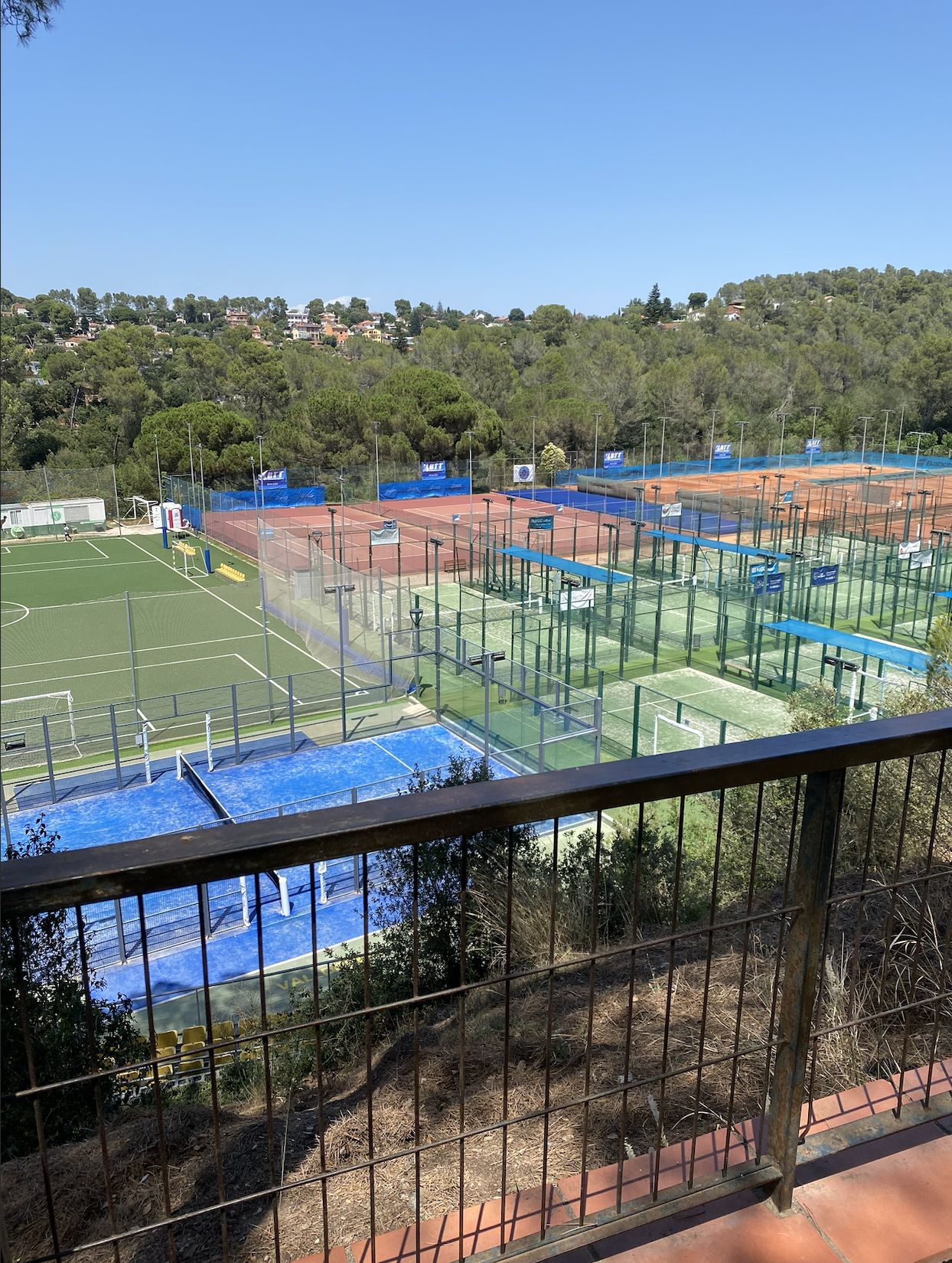 A view from a balcony showing multiple tennis courts with orange clay and green surfaces, surrounded by fences and trees, with houses on a hillside in the background under a clear blue sky.