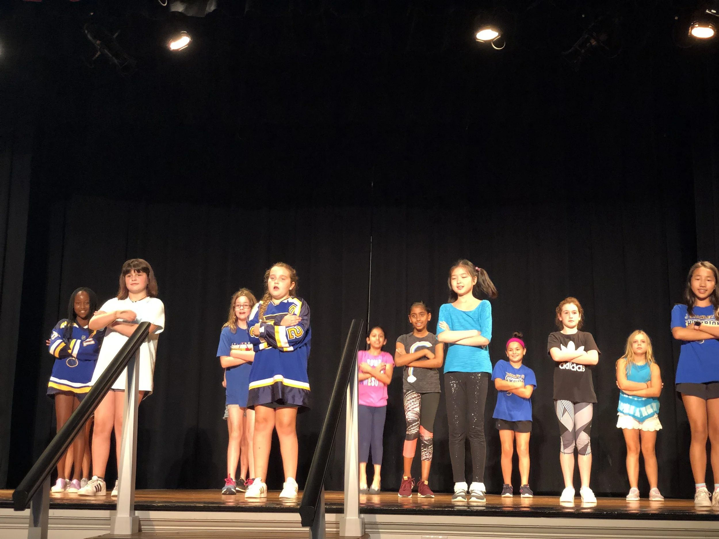 A group of children standing on stage with their arms crossed during a performance or rehearsal.