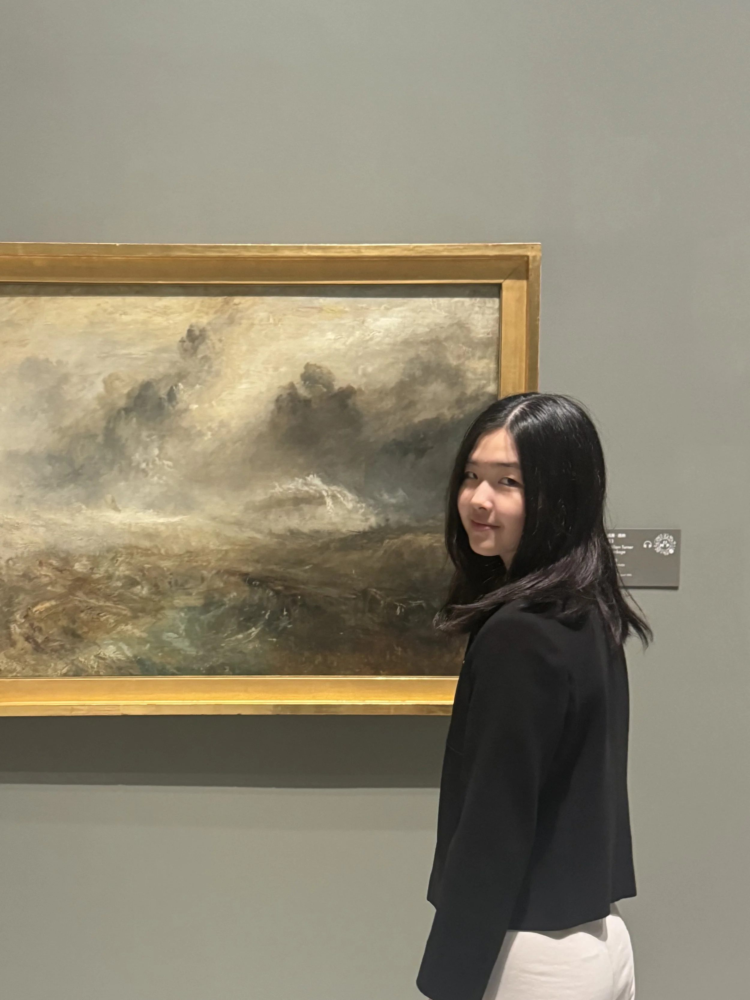 A young woman with black hair in a black blazer and white pants standing at an art museum, viewing a landscape painting with dark clouds and a stormy scene.