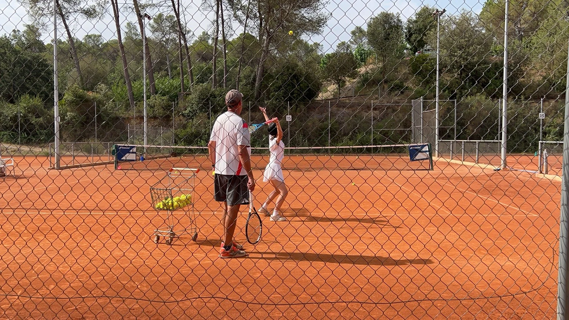 A man and a young girl playing tennis on an outdoor clay tennis court, surrounded by a metal chain-link fence, with trees and bushes in the background.