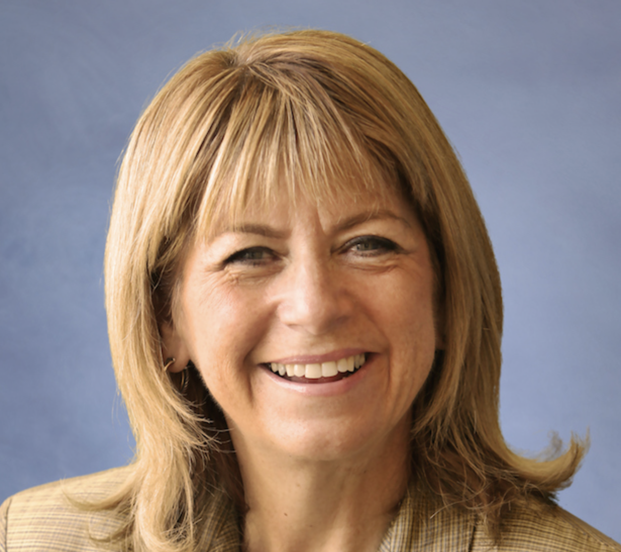 A smiling woman with shoulder-length blonde hair, wearing a checkered blazer, against a blue background.